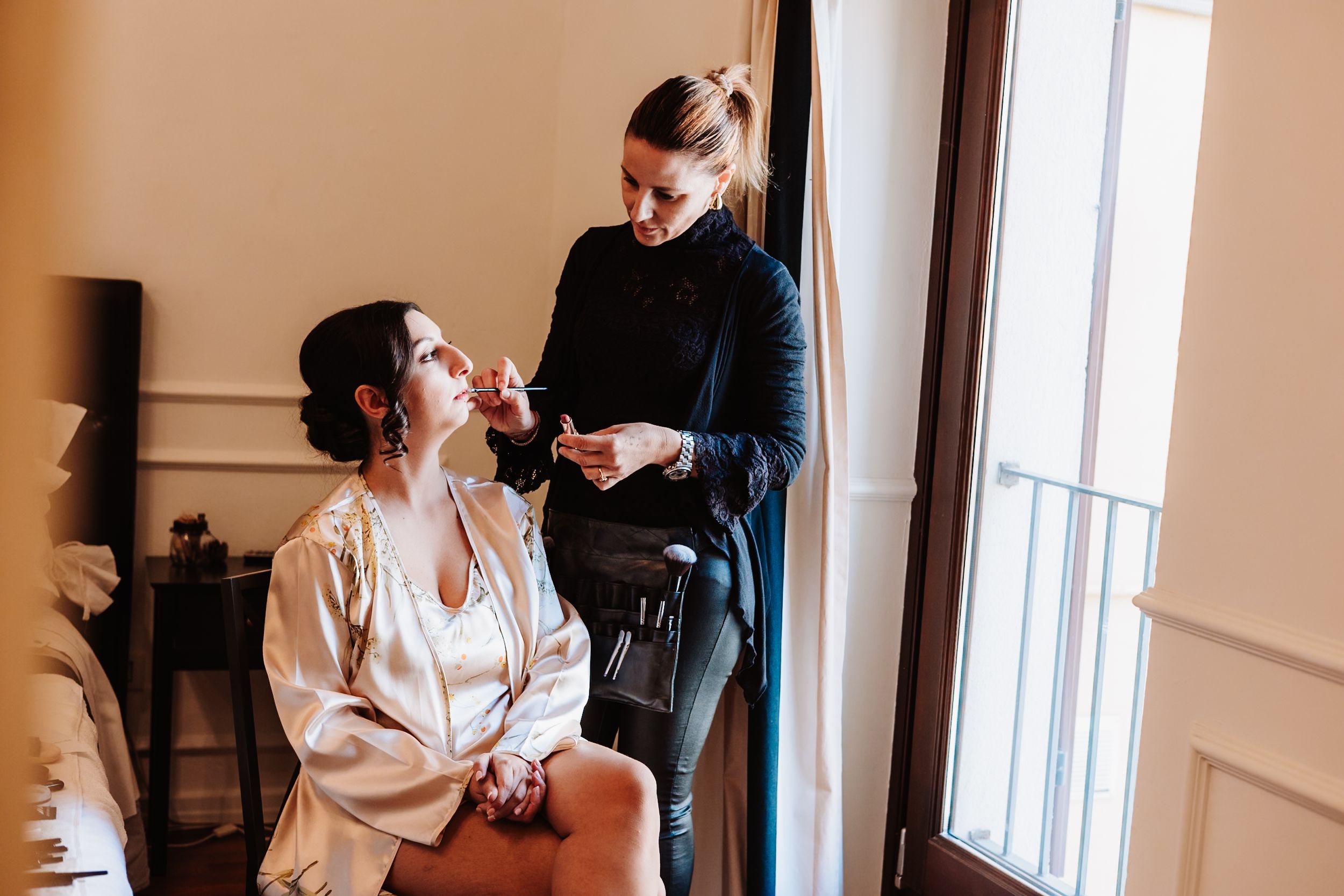 A bride and bridesmaids getting ready in a sunlit room with white robes and wedding preparations underway.