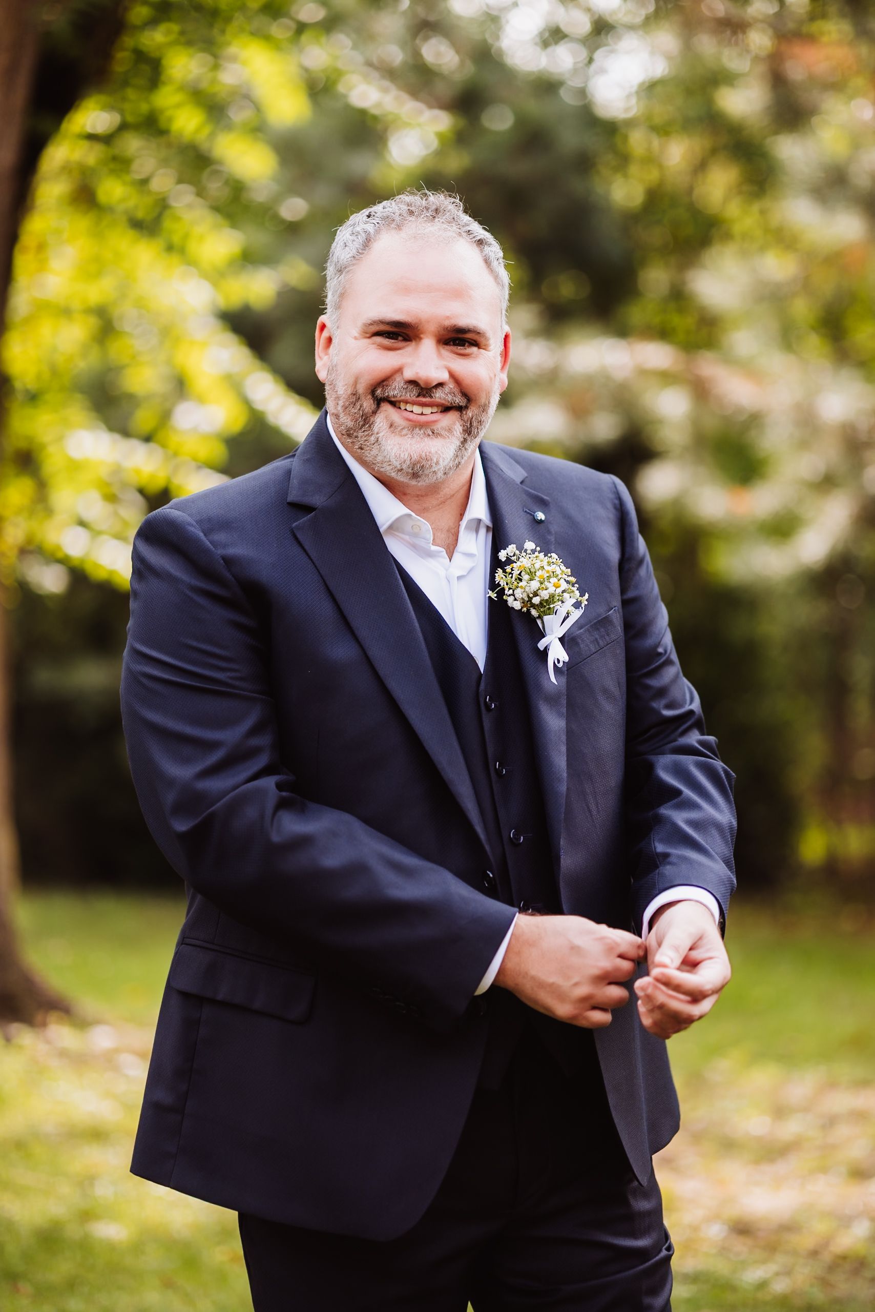 A person in a dark suit adjusts their boutonniere while smiling outdoors in a garden setting.