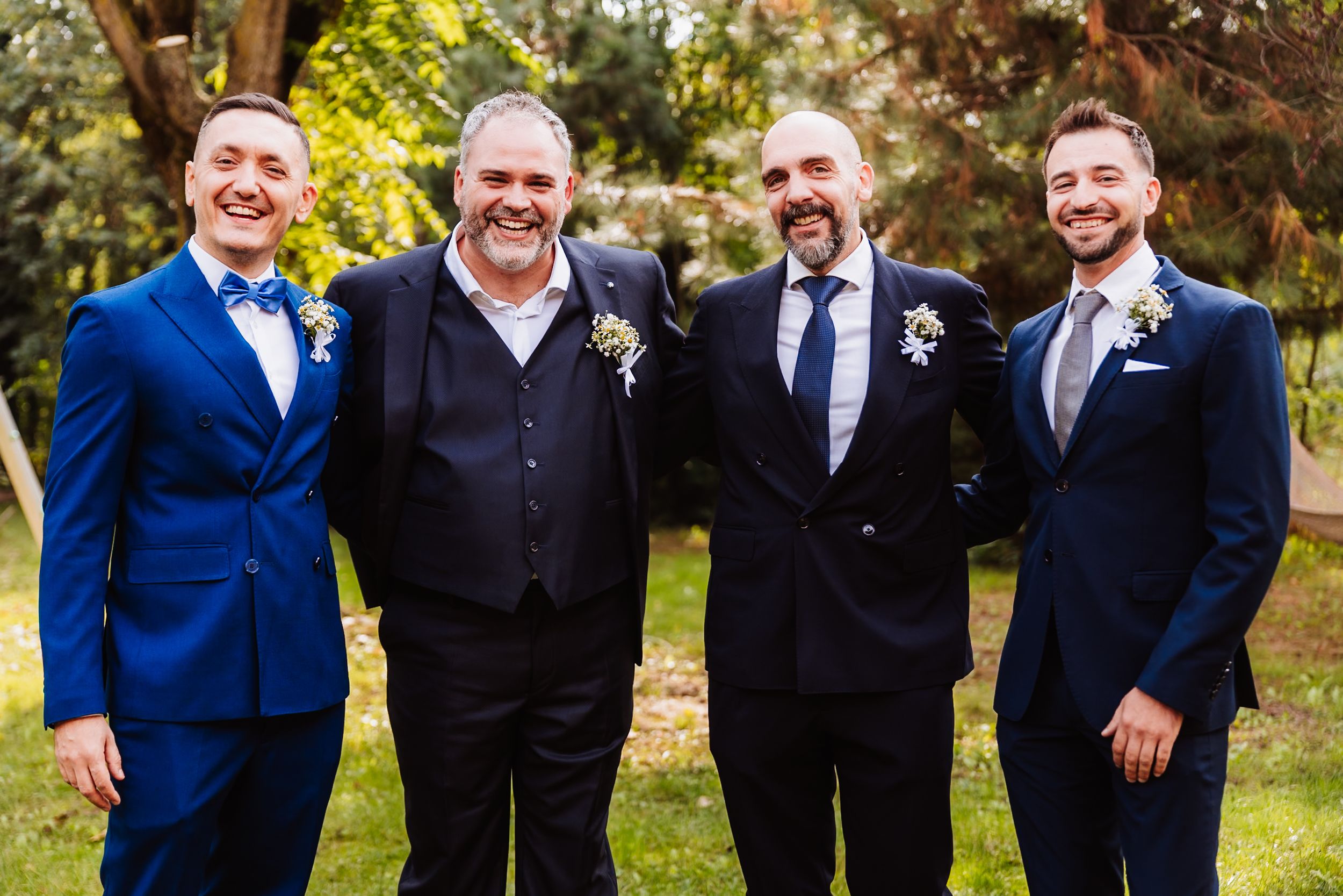 Four men in formal attire stand together smiling outdoors with blue suits and boutonnieres at a wedding celebration.