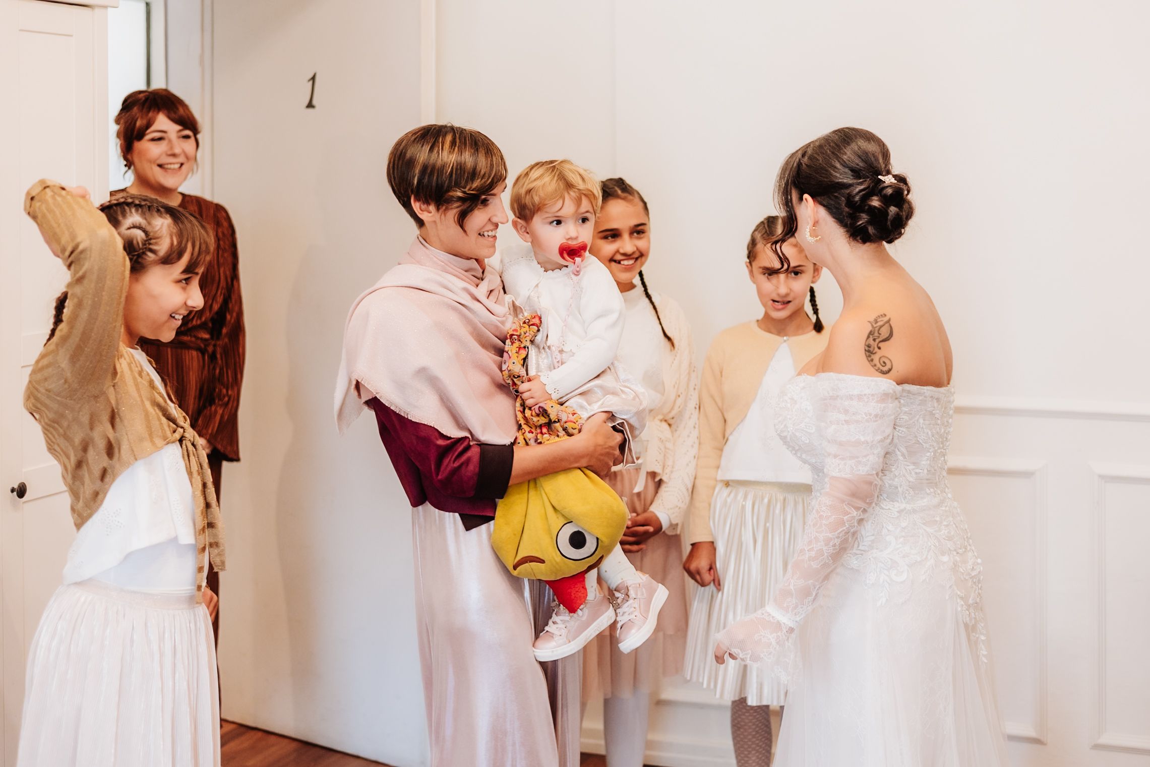 A group of wedding party members in white dresses dance and celebrate together in a bright indoor venue.