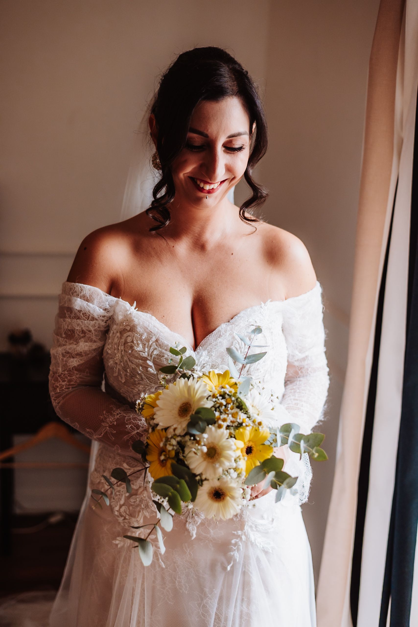 A bride in an off-shoulder lace wedding dress holds a delicate bouquet of white and yellow flowers.