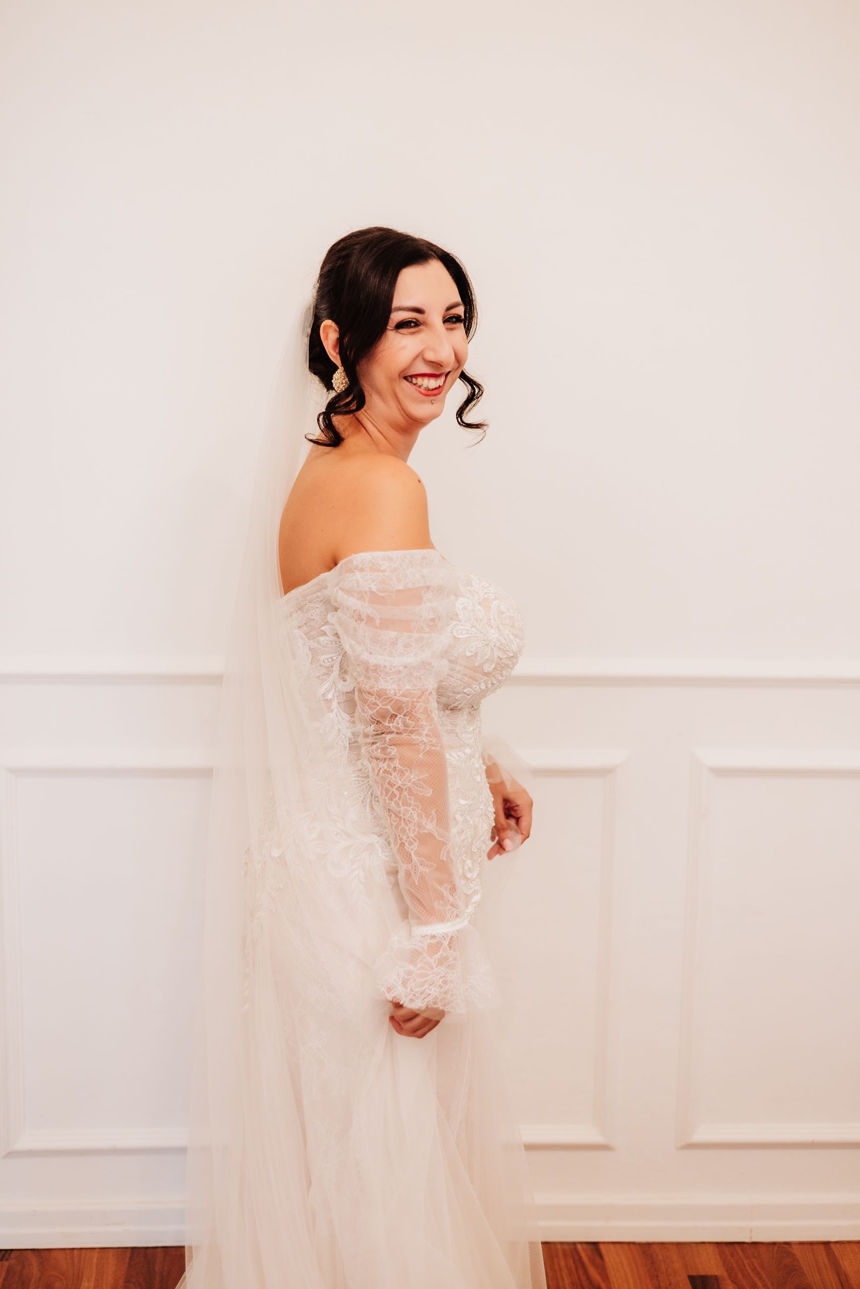 A bride wearing an ethereal off-shoulder lace wedding gown poses against a white wall in a series of elegant portraits.