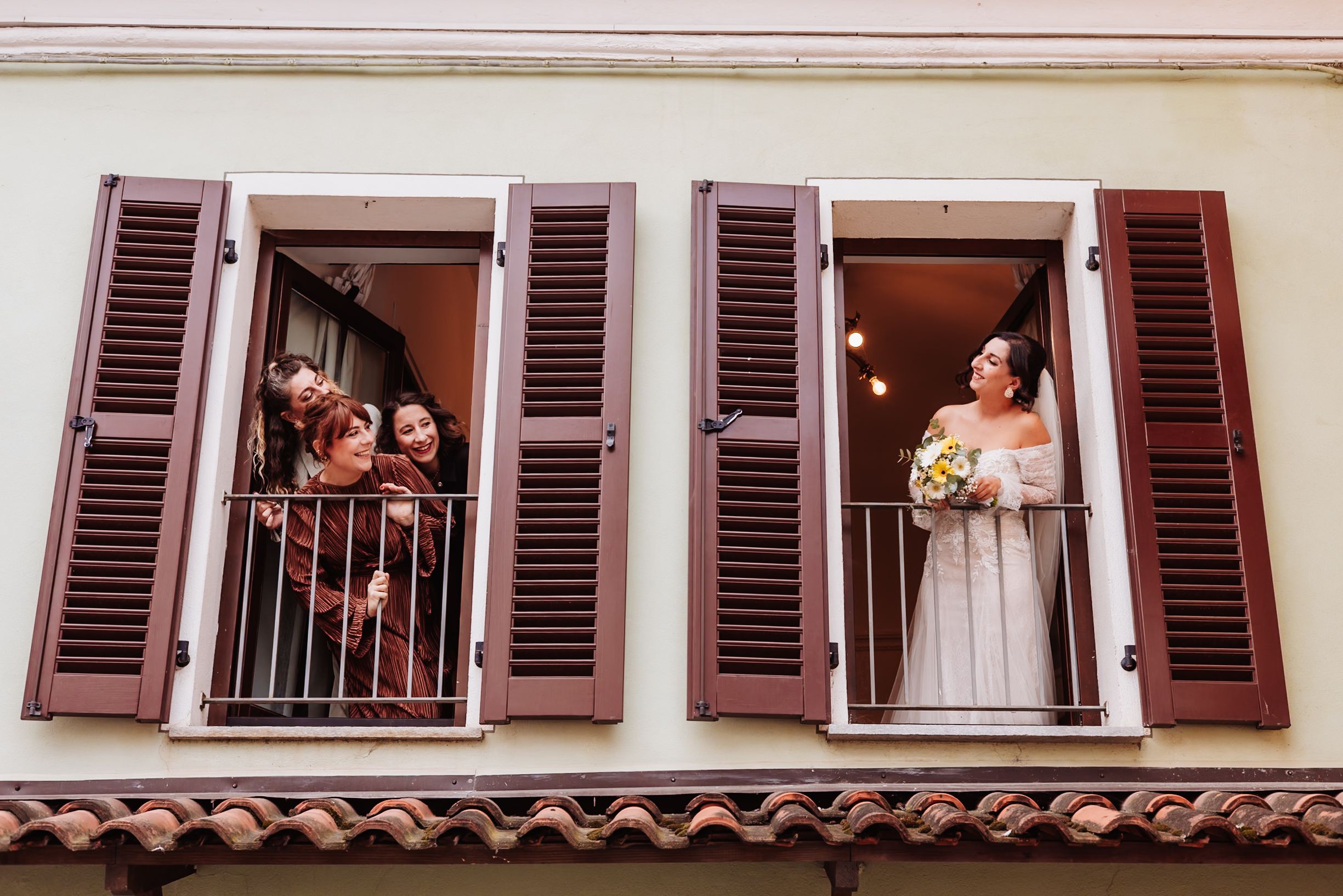 Couples lean out of windows with brown shutters in a romantic scene at a Mediterranean style building.