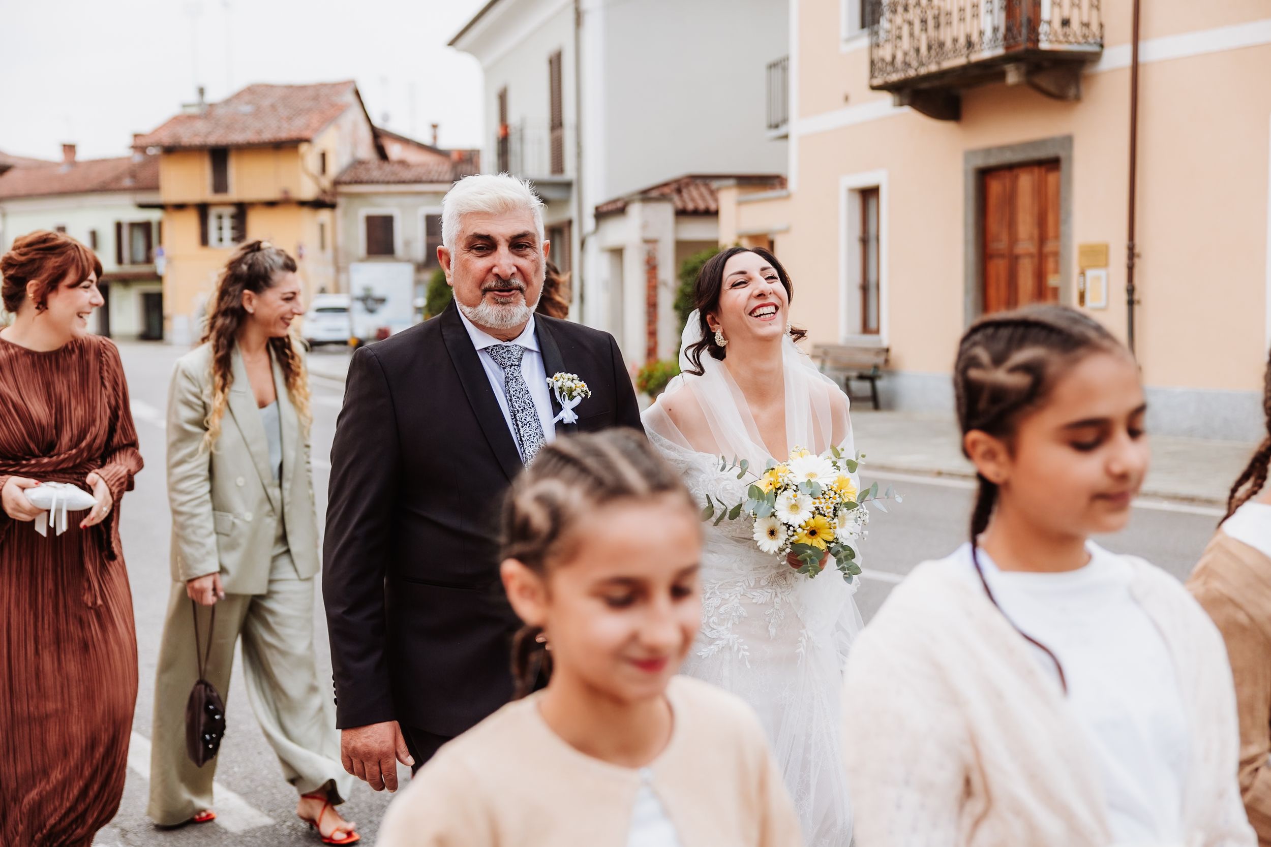 A wedding party walks together in a line outside a peach-colored building on a sunny day.