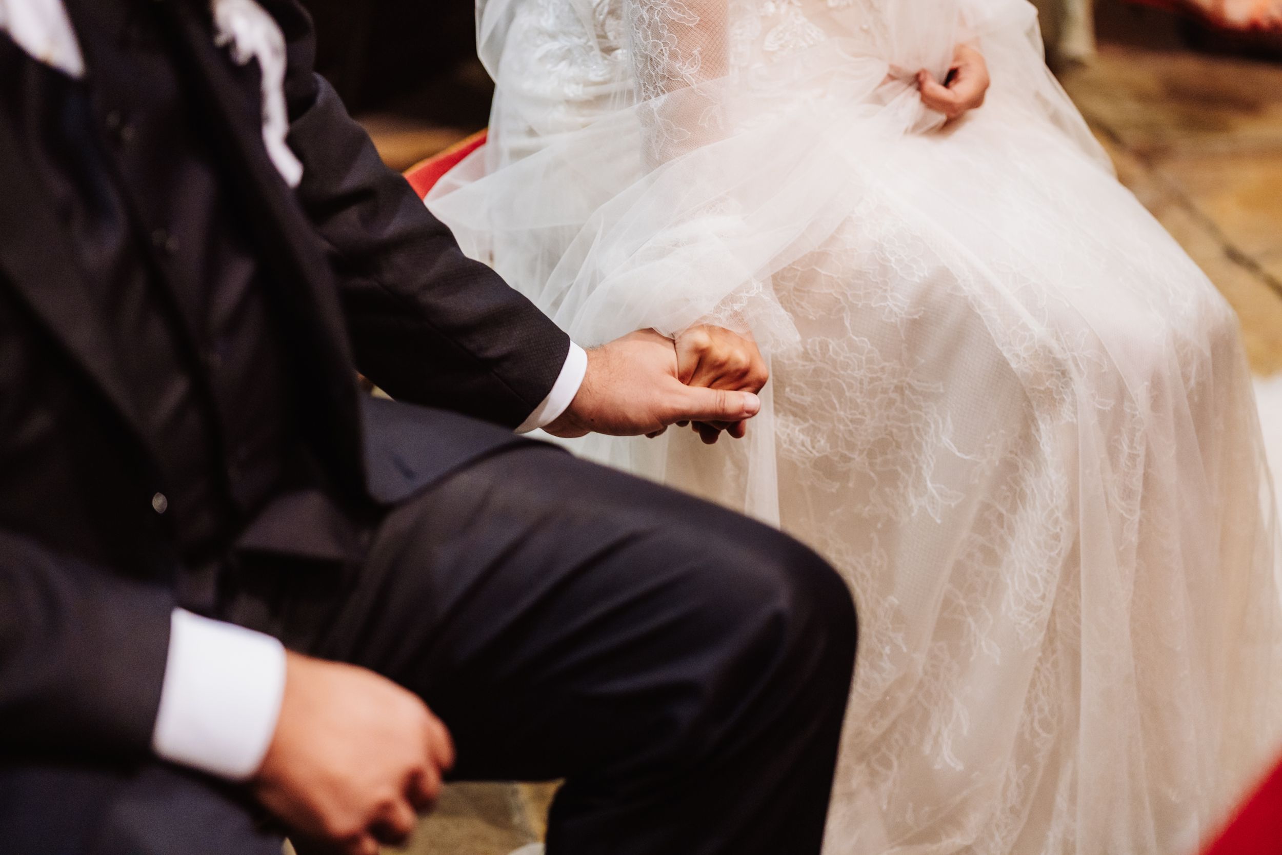 A close-up shot of a couple holding hands during their wedding ceremony, with white bridal gown and black suit visible.