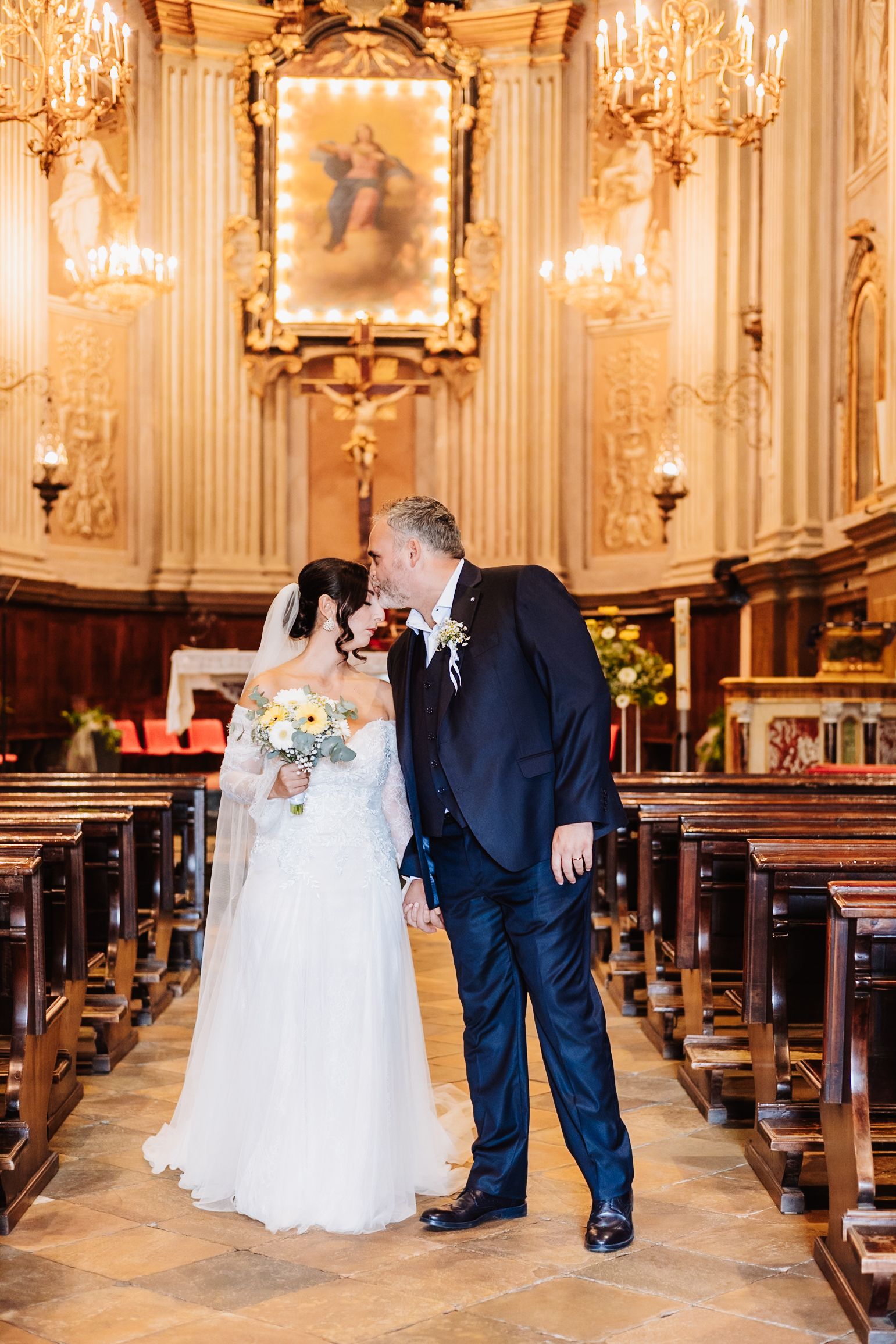 Elegant wedding ceremony in an ornate baroque church with golden chandeliers and wooden pews.