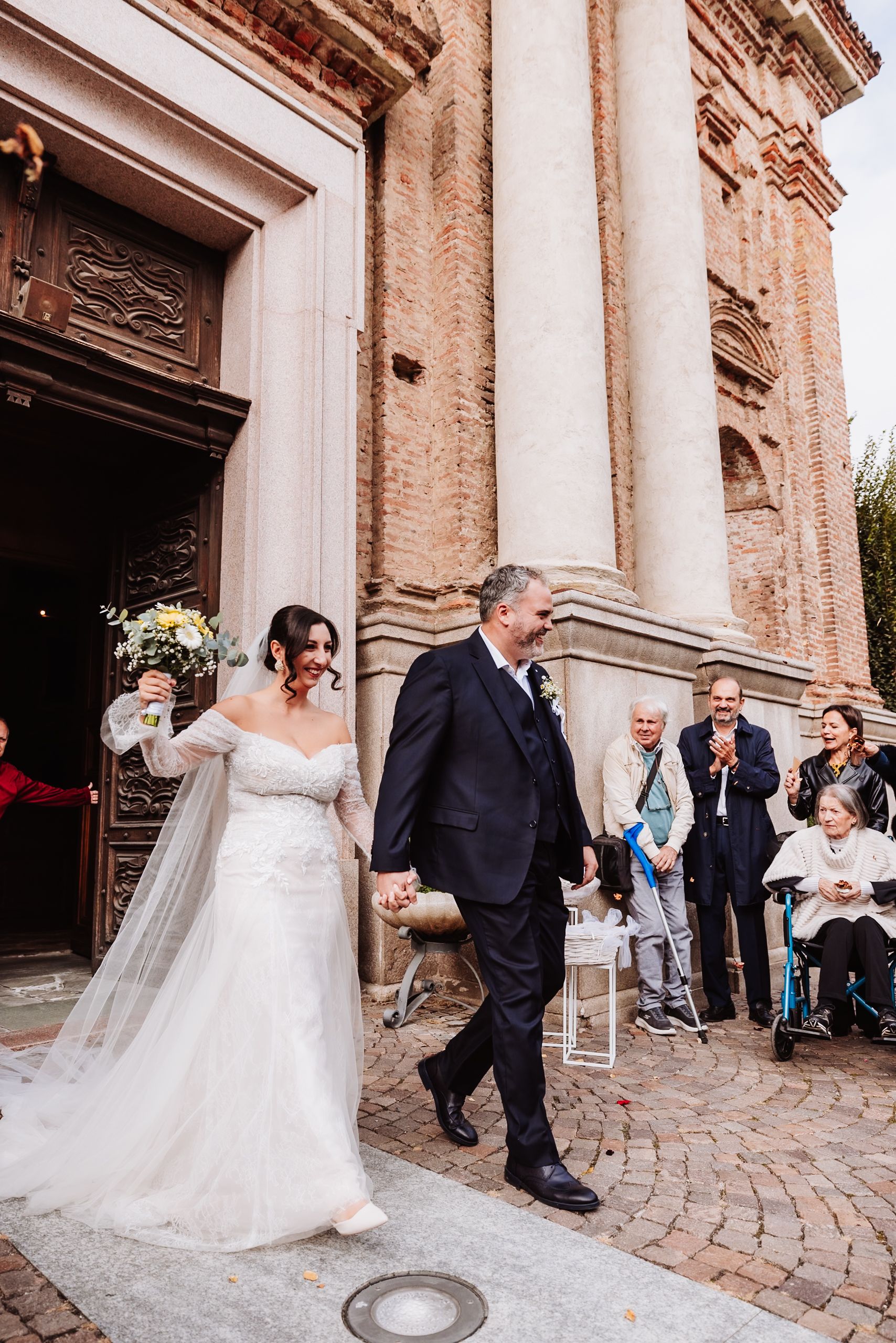 Newlywed couple in formal attire walks together outside a historic church with stone columns and cobblestone path.