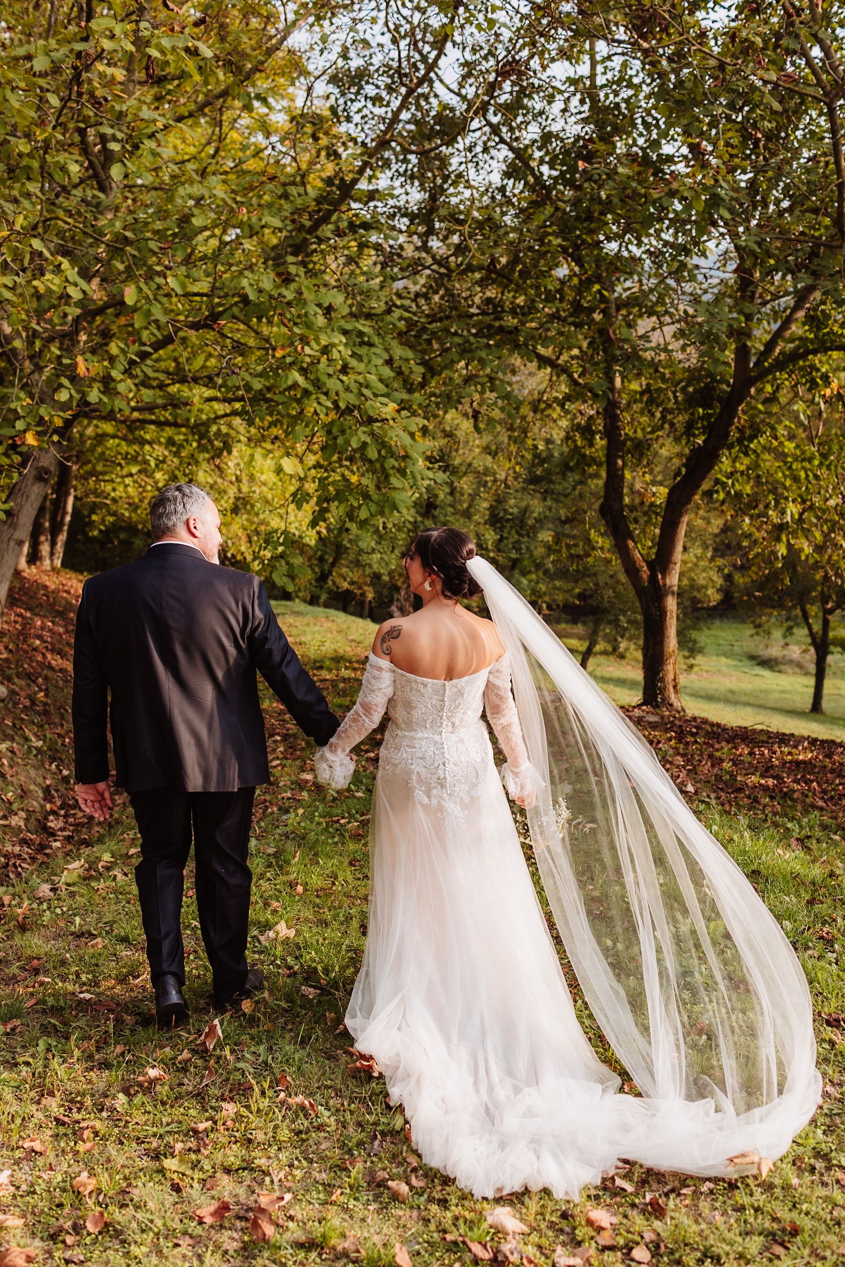 A couple in wedding attire walks through a scenic autumn orchard with golden leaves and sunlight filtering through trees.