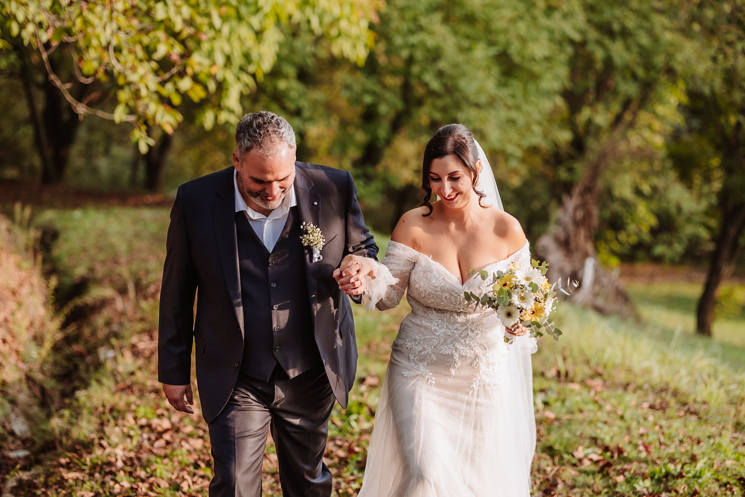 Couple in wedding attire walking through a scenic autumn garden path with golden foliage.