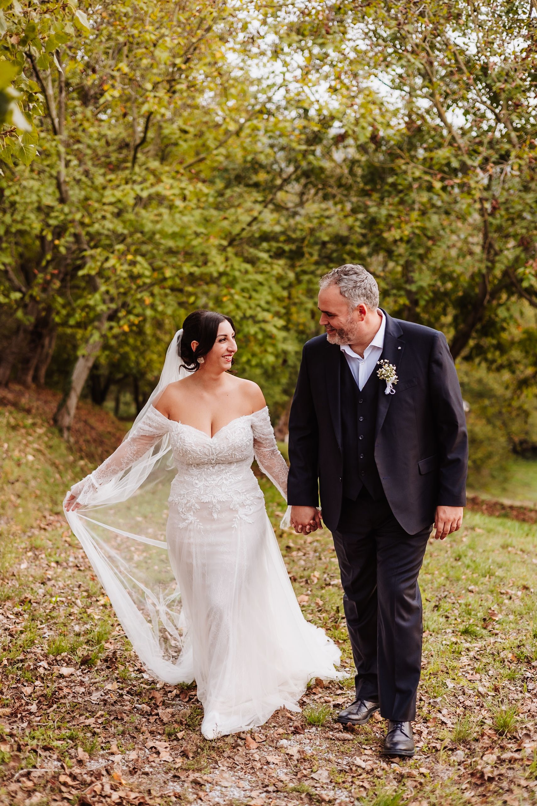 A romantic wedding couple walks through autumn leaves as the bride's lace veil flows in the breeze.