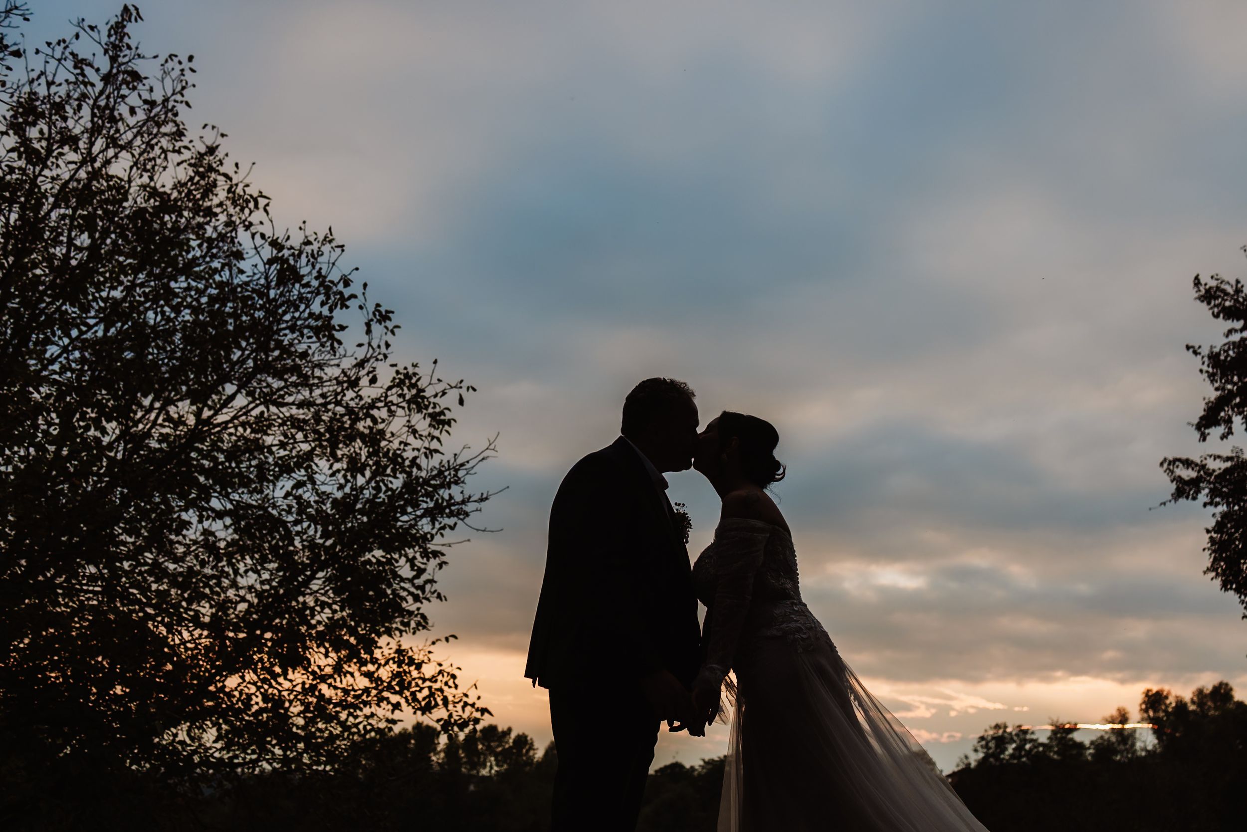 A silhouetted couple shares an intimate moment against a dramatic sunset sky with trees framing the scene.