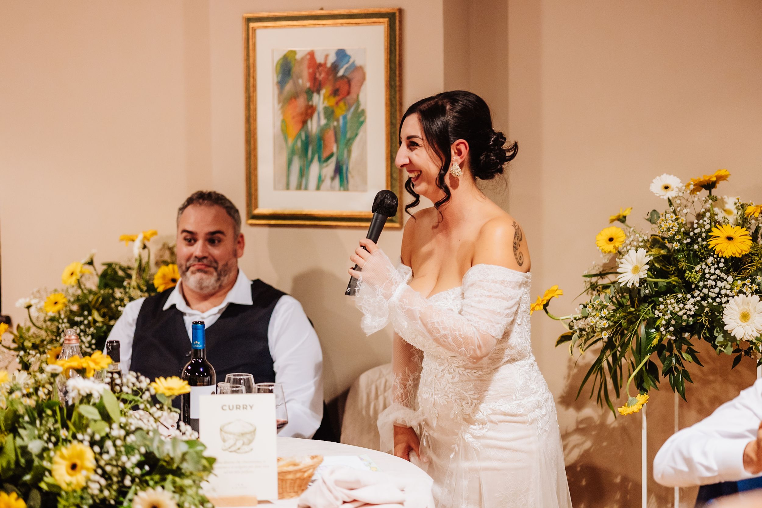 A joyful wedding reception scene with yellow floral decorations and speakers at the head table during speeches.
