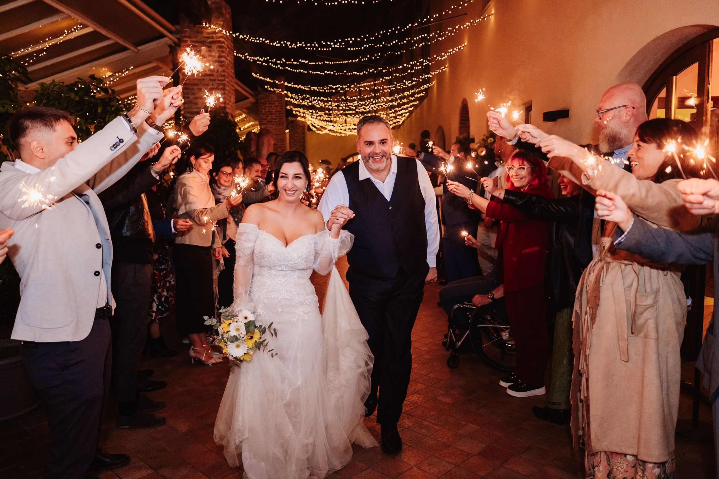Wedding couple walks down aisle while guests cheer and wave sparklers during evening reception celebration.