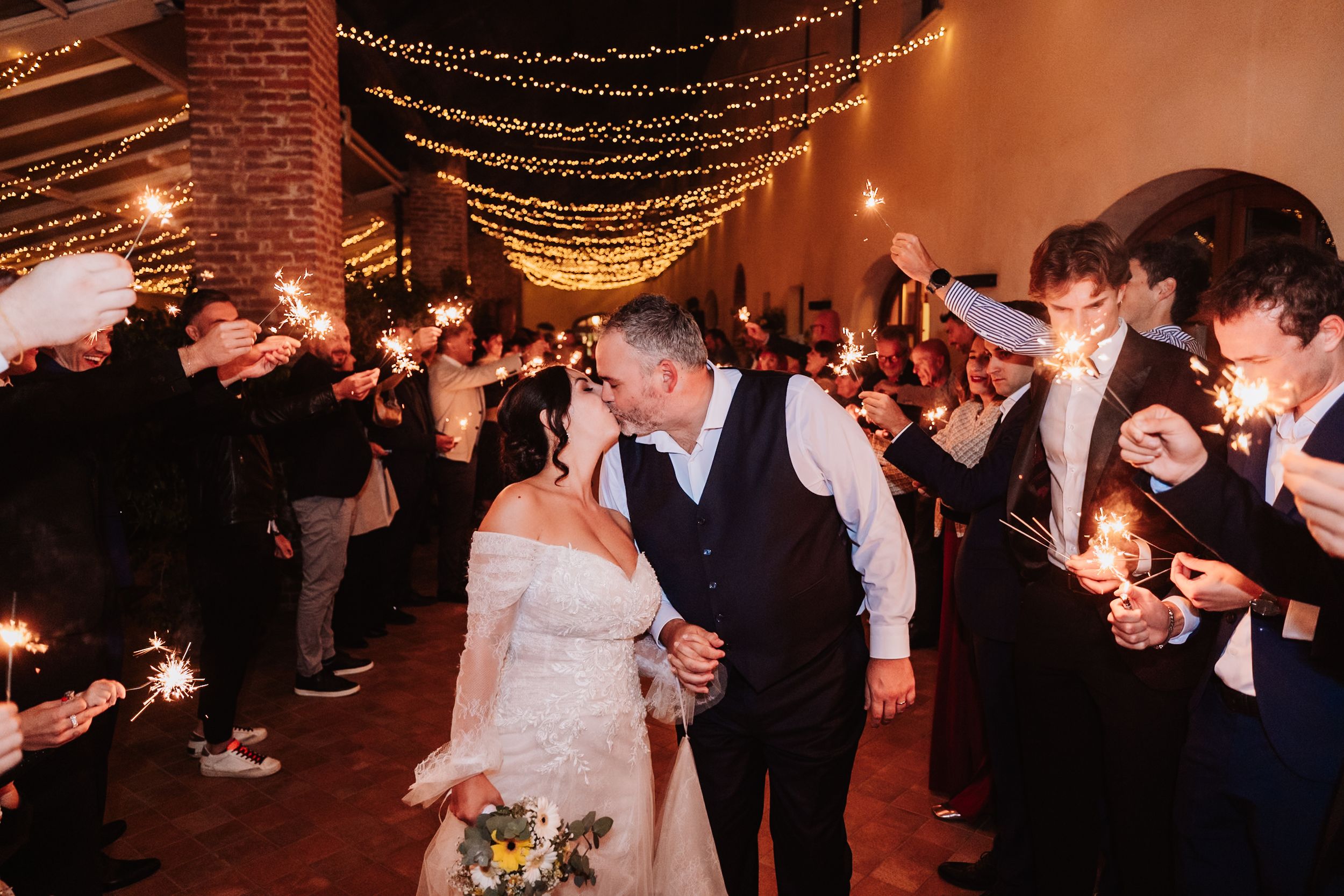 Wedding guests dance together under warm string lights at an indoor reception with brick walls.