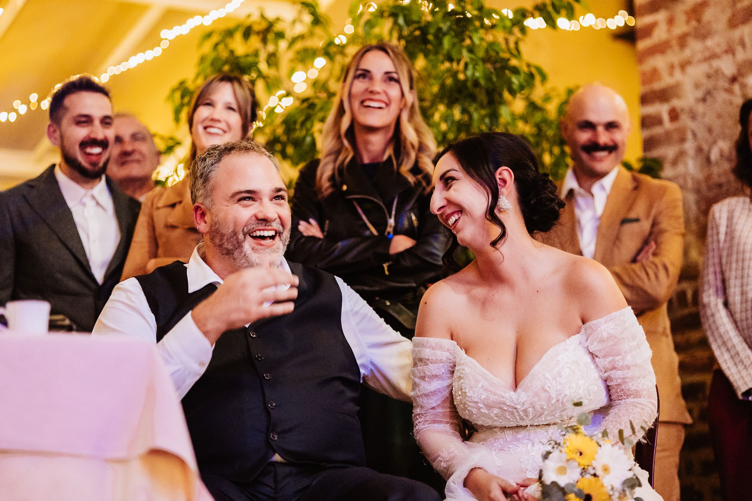 A joyful wedding celebration with people laughing together during a reception in a warmly lit venue.