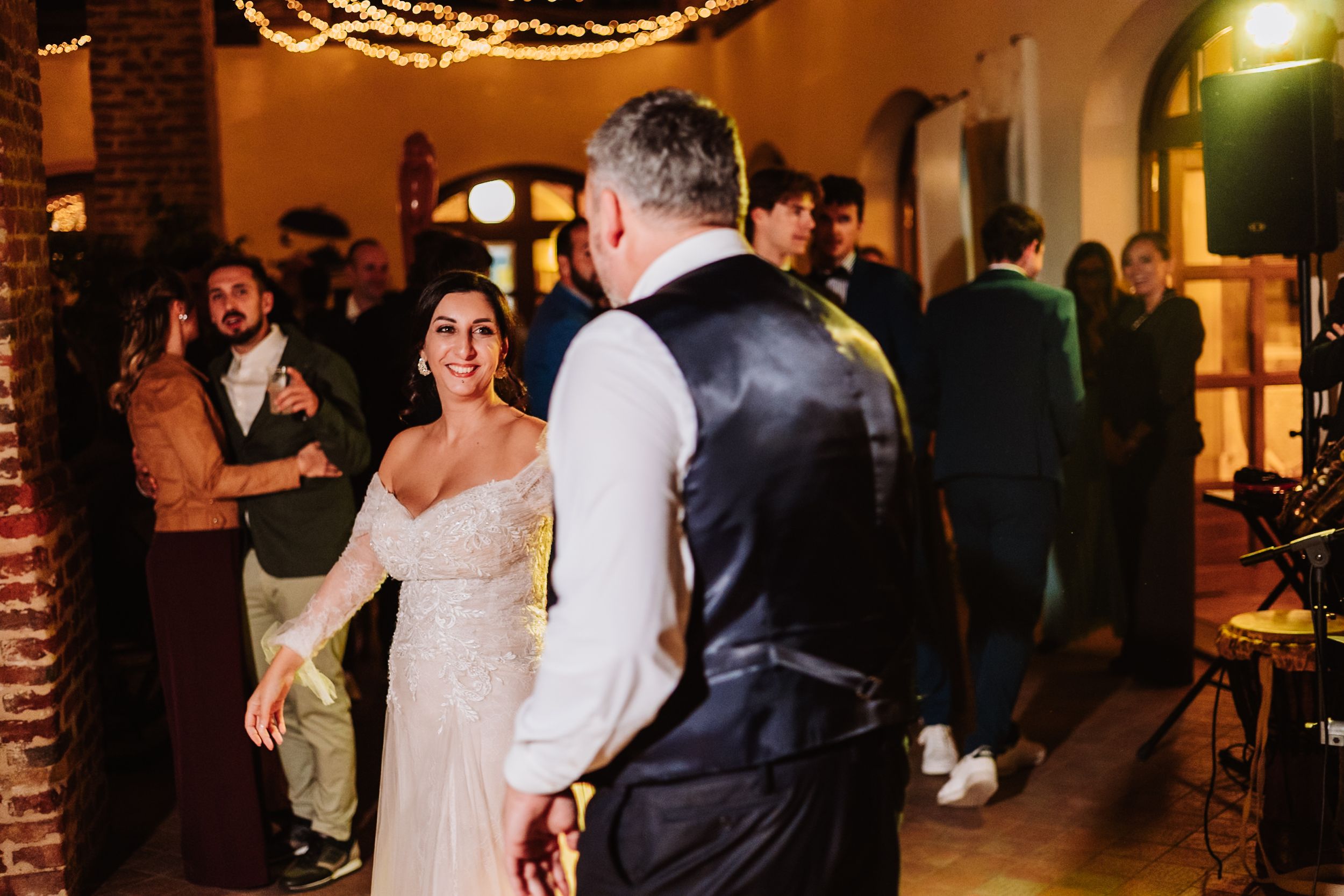 Wedding guests dance and celebrate in an elegant reception venue with warm lighting and brick columns.