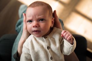 A baby in a cream-colored sweater sits in natural light while making various expressions in a photo series.