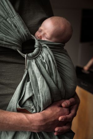 A sleeping baby nestled in a sage green fabric baby carrier wrap, supported by caring hands.