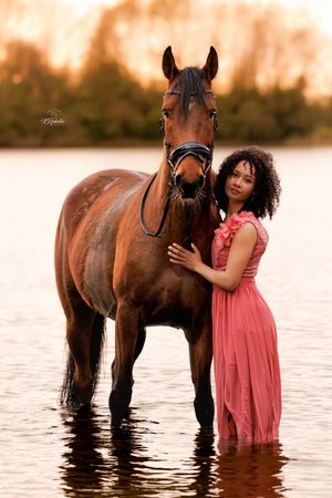 Woman in pink dress stands with brown horse in water at golden sunset.