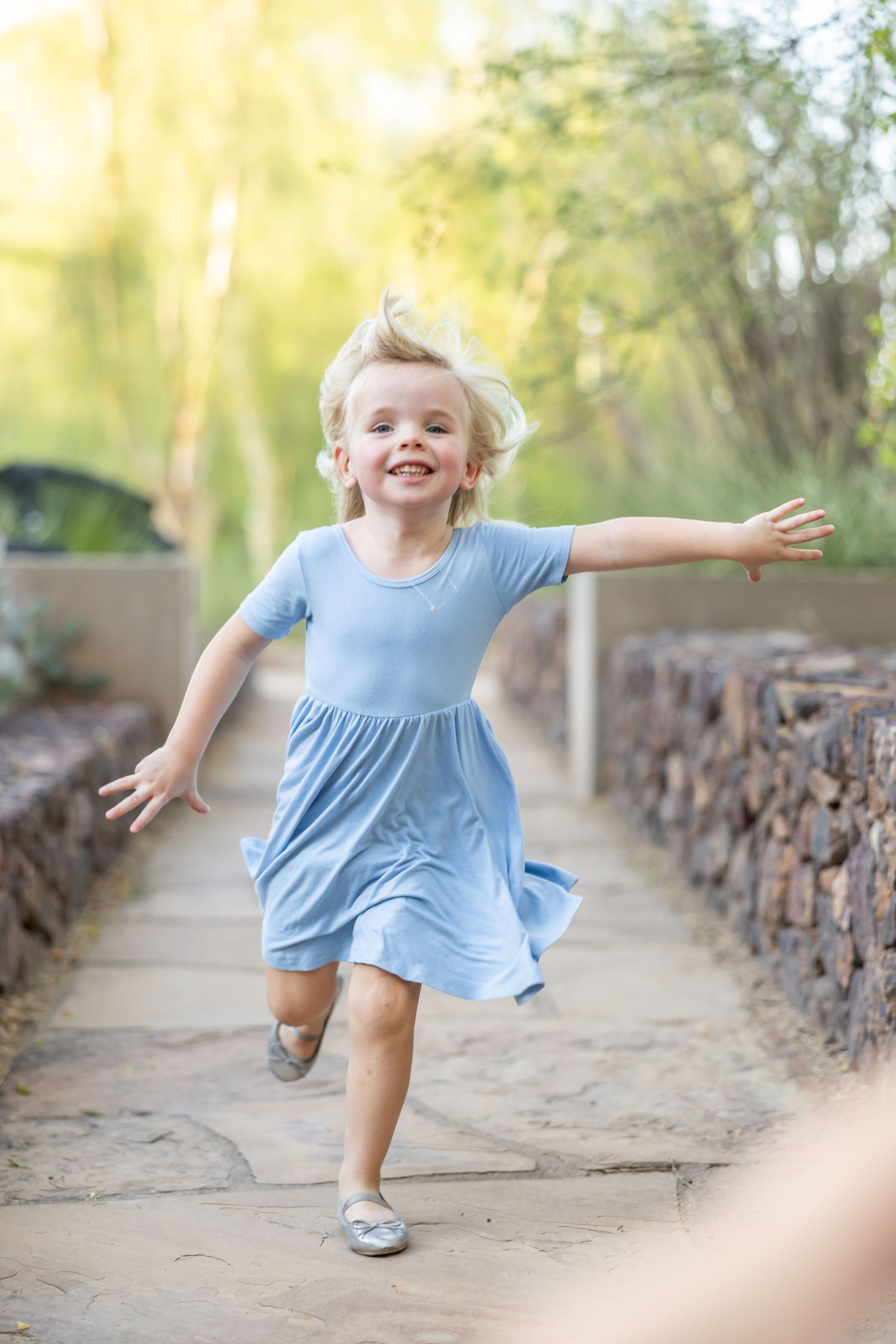 A young child in a light blue dress twirls and dances joyfully on a dirt path lined with stone walls.