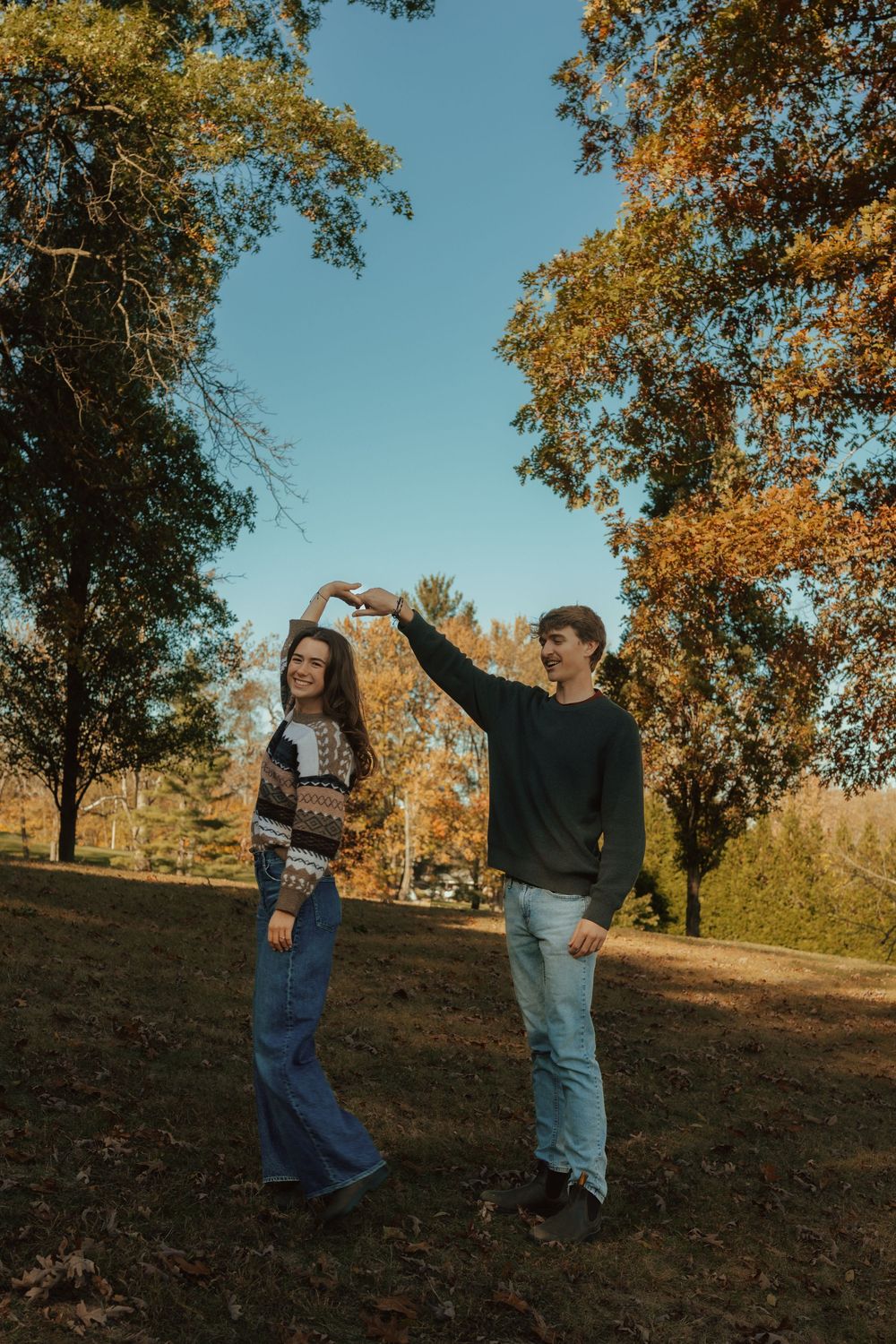 Two children play together on a grassy hillside during autumn with golden leaves on trees in the background.