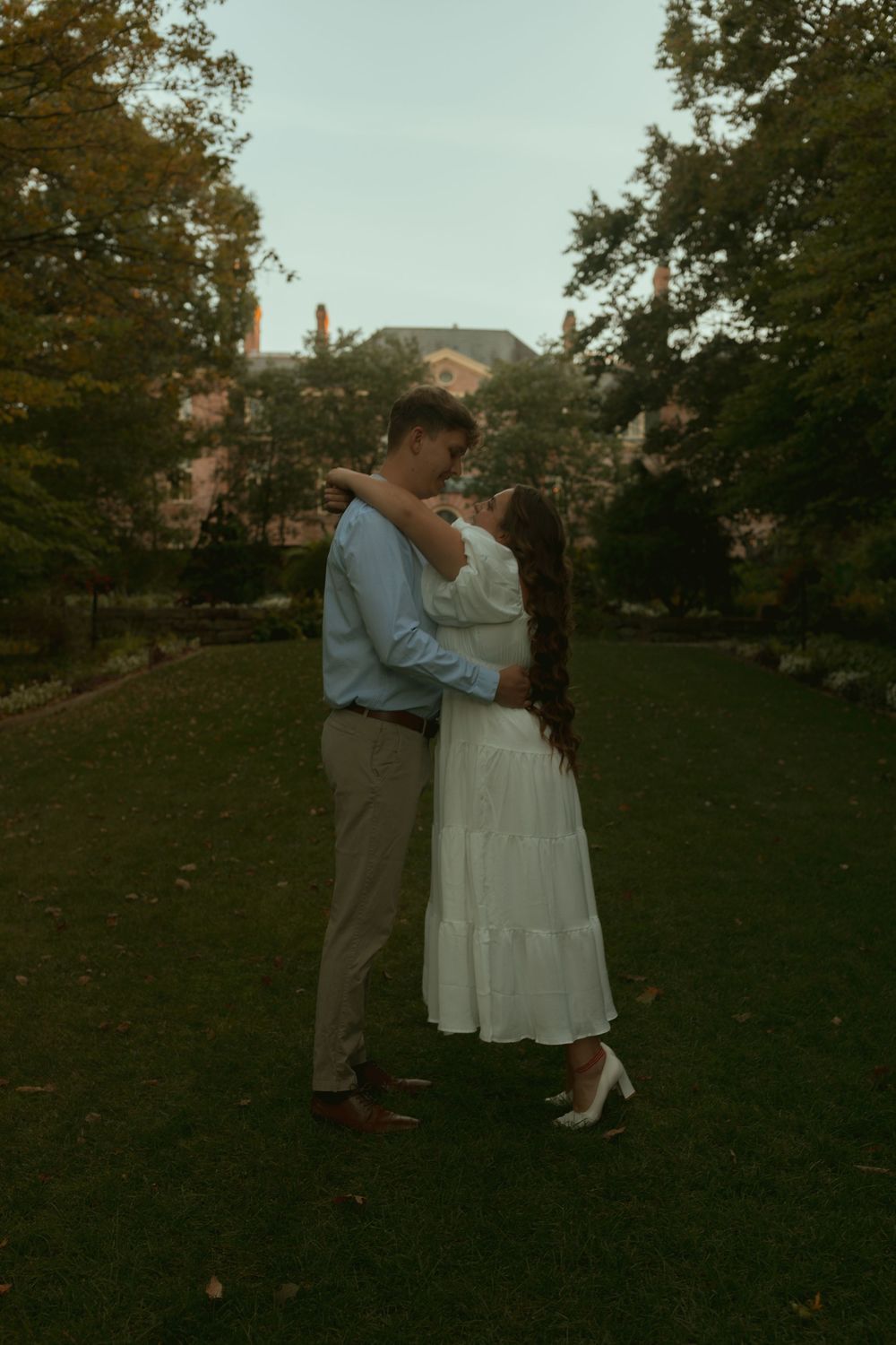 A couple shares a romantic embrace on a grassy lawn at dusk with trees and houses visible in the background.
