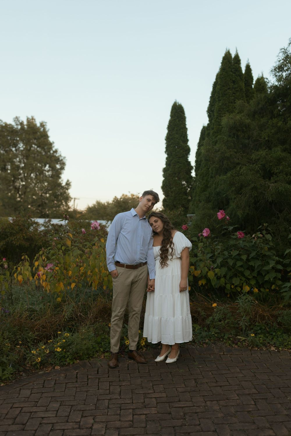 A couple stands together on a brick path in a garden with tall cypress trees and flowers at sunset.