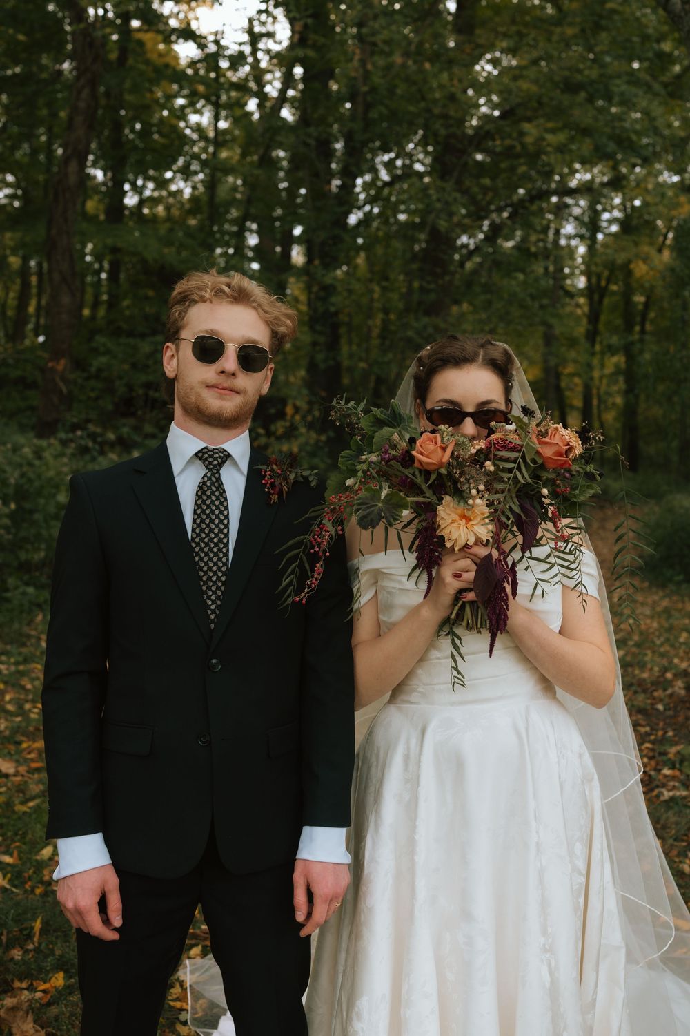 A playful wedding couple in sunglasses poses together in a wooded outdoor setting during autumn.