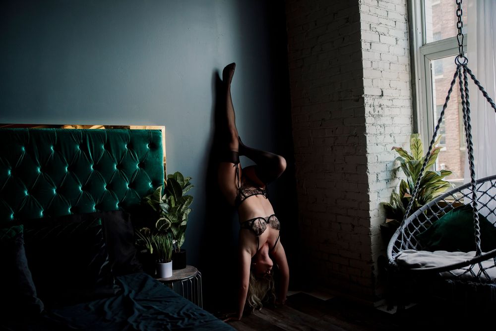 Woman in lingerie performing a handstand against a dark teal wall near a window.