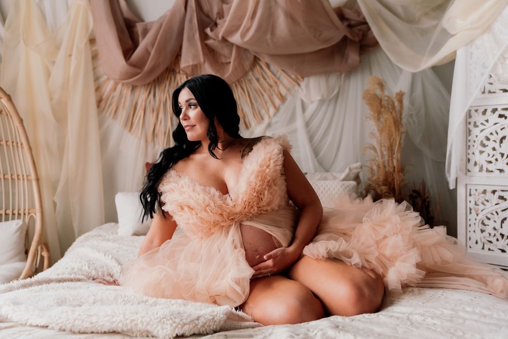 Pregnant woman in a peach tulle robe sitting on a bed in a boho studio.