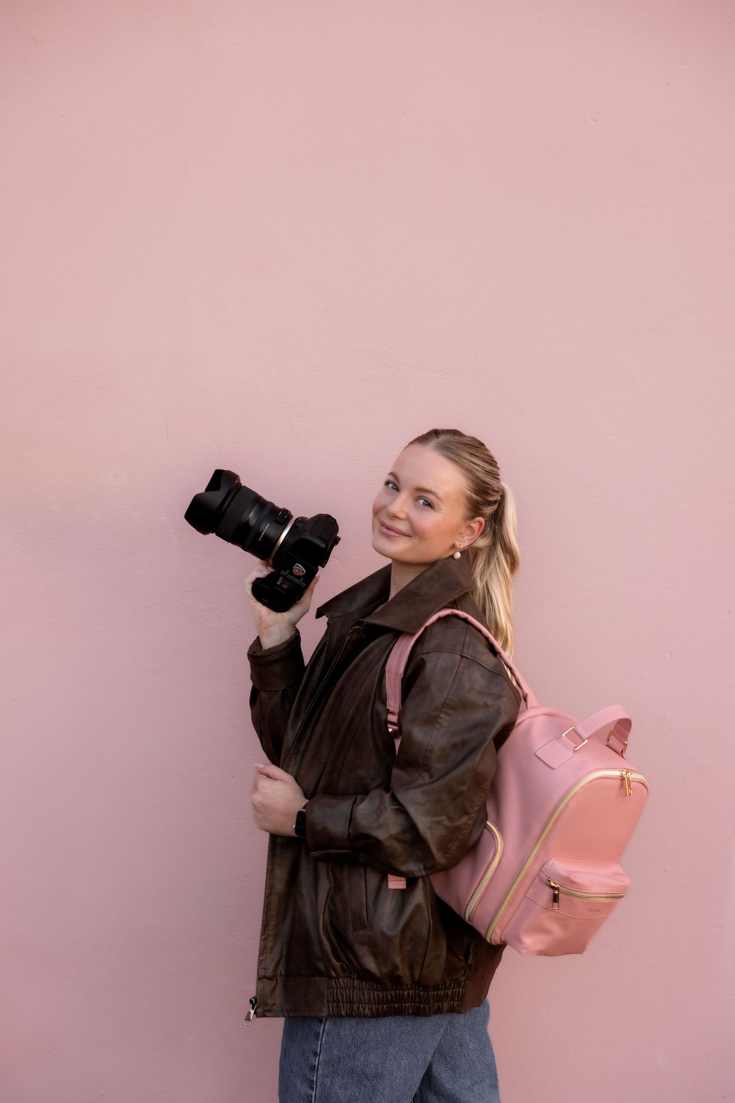 Young blonde woman holding a DSLR camera, wearing a brown jacket and pink backpack, posing against a pink wall.
