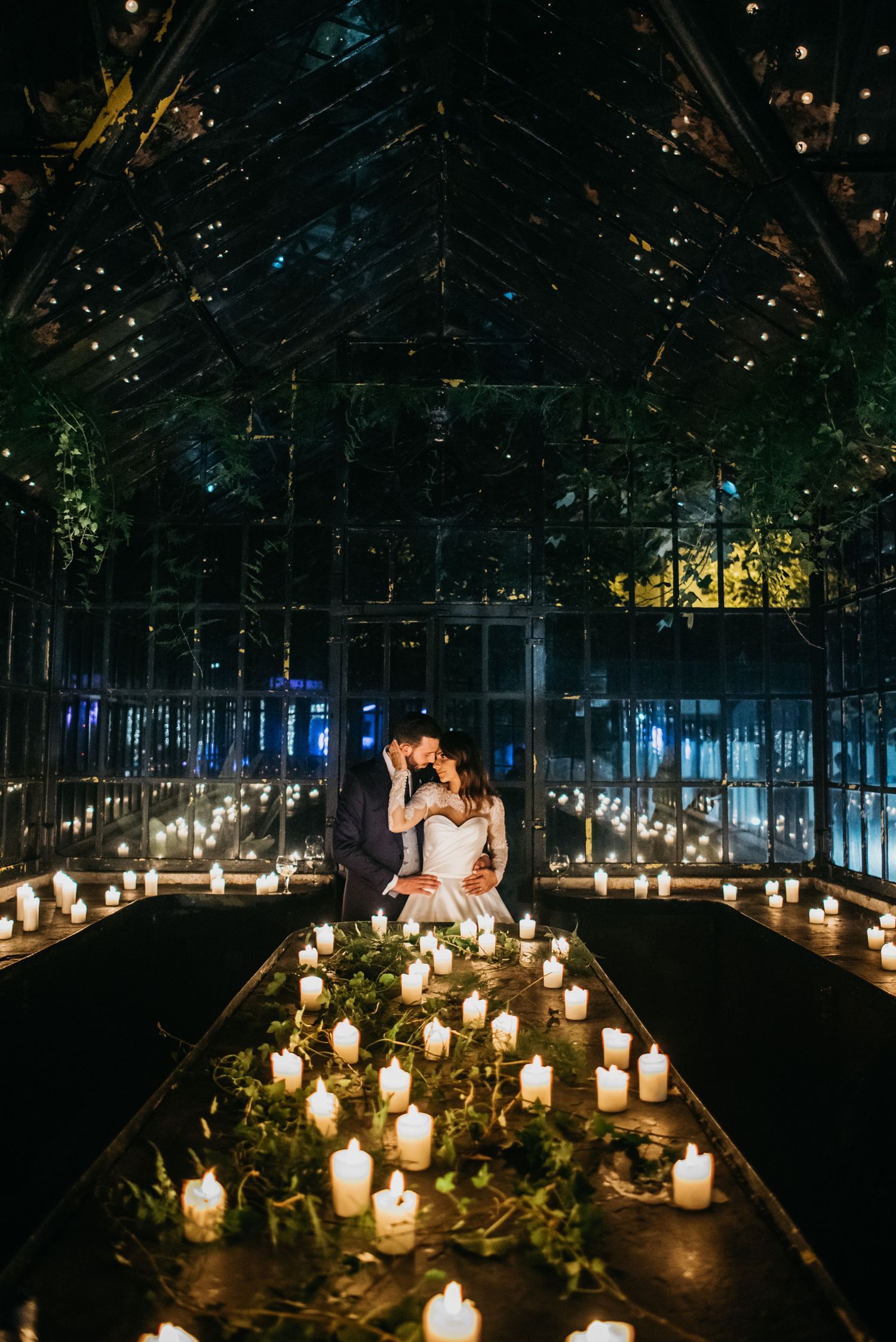 A romantic candlelit wedding scene in a glass greenhouse with glowing candles lining the aisle at night.