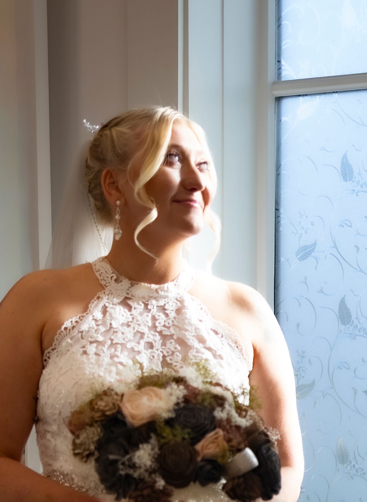 A bride in a white lace wedding dress poses by a window with soft natural lighting and holds a floral bouquet.