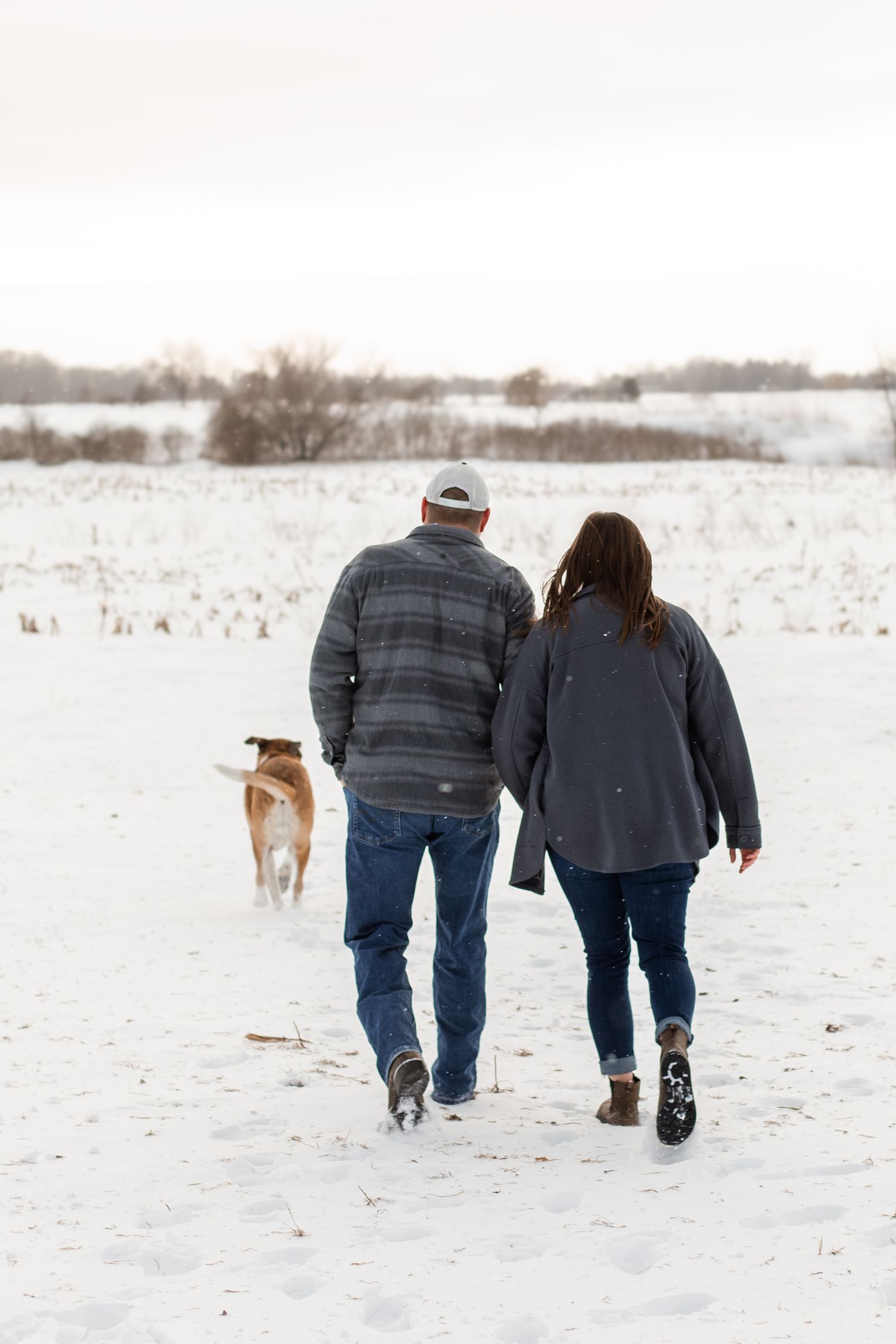 A couple walks together through a snowy winter landscape with their dog following behind them.
