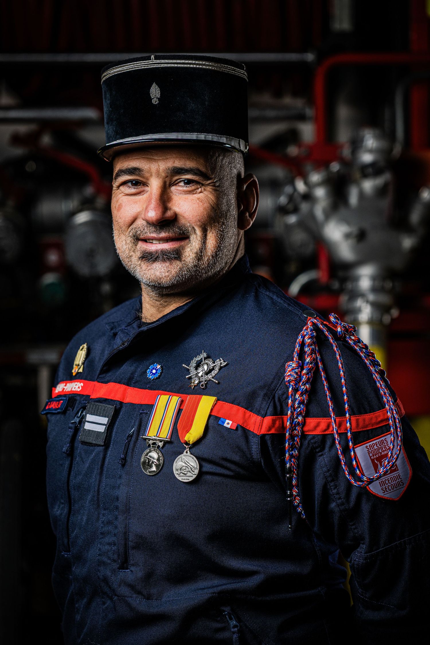 French firefighter in traditional navy blue uniform with medals and kepi hat stands in front of blurred fire station background.