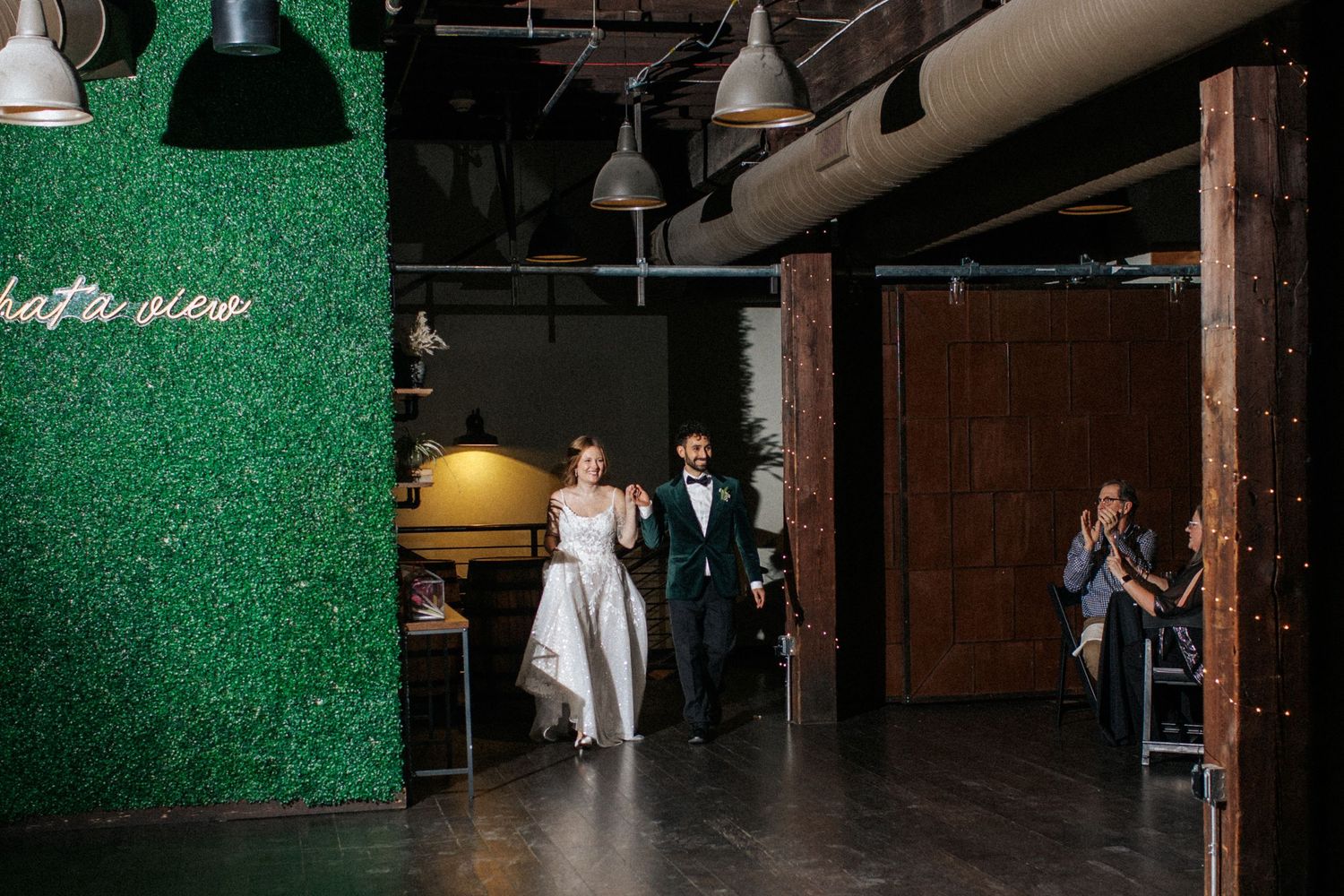 Wedding reception guests dancing and celebrating in an industrial-style venue with exposed brick walls and hanging lights.