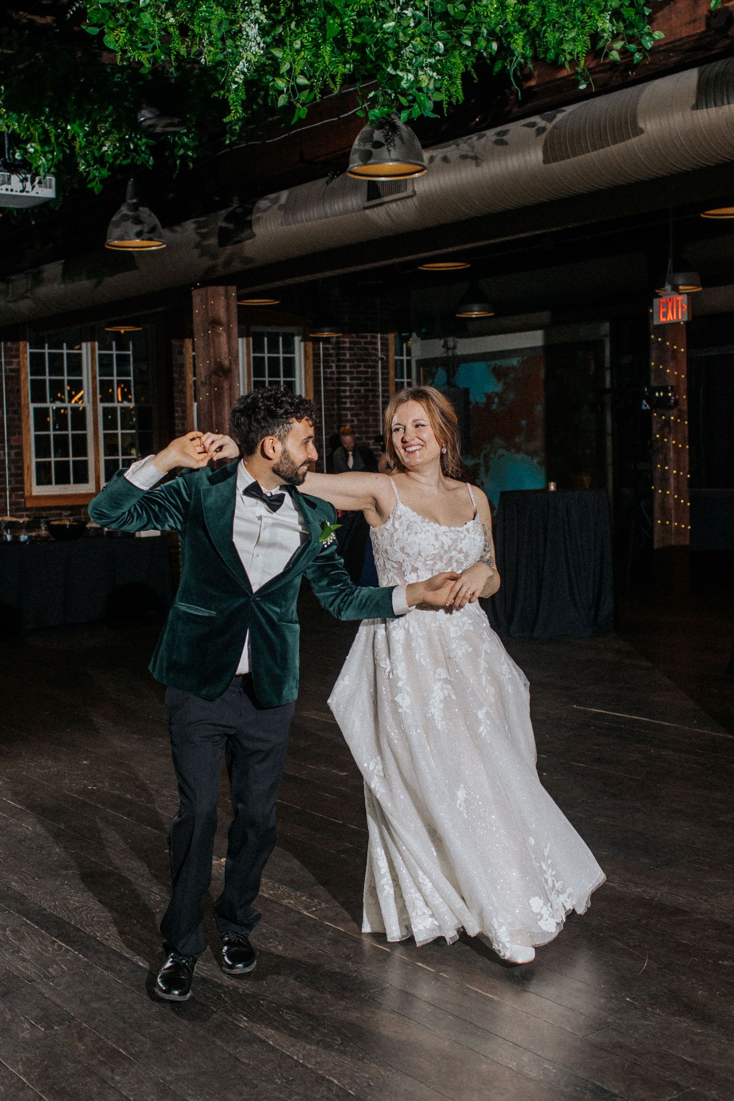 Wedding couple performs a choreographed dance routine on a wooden floor under hanging greenery.