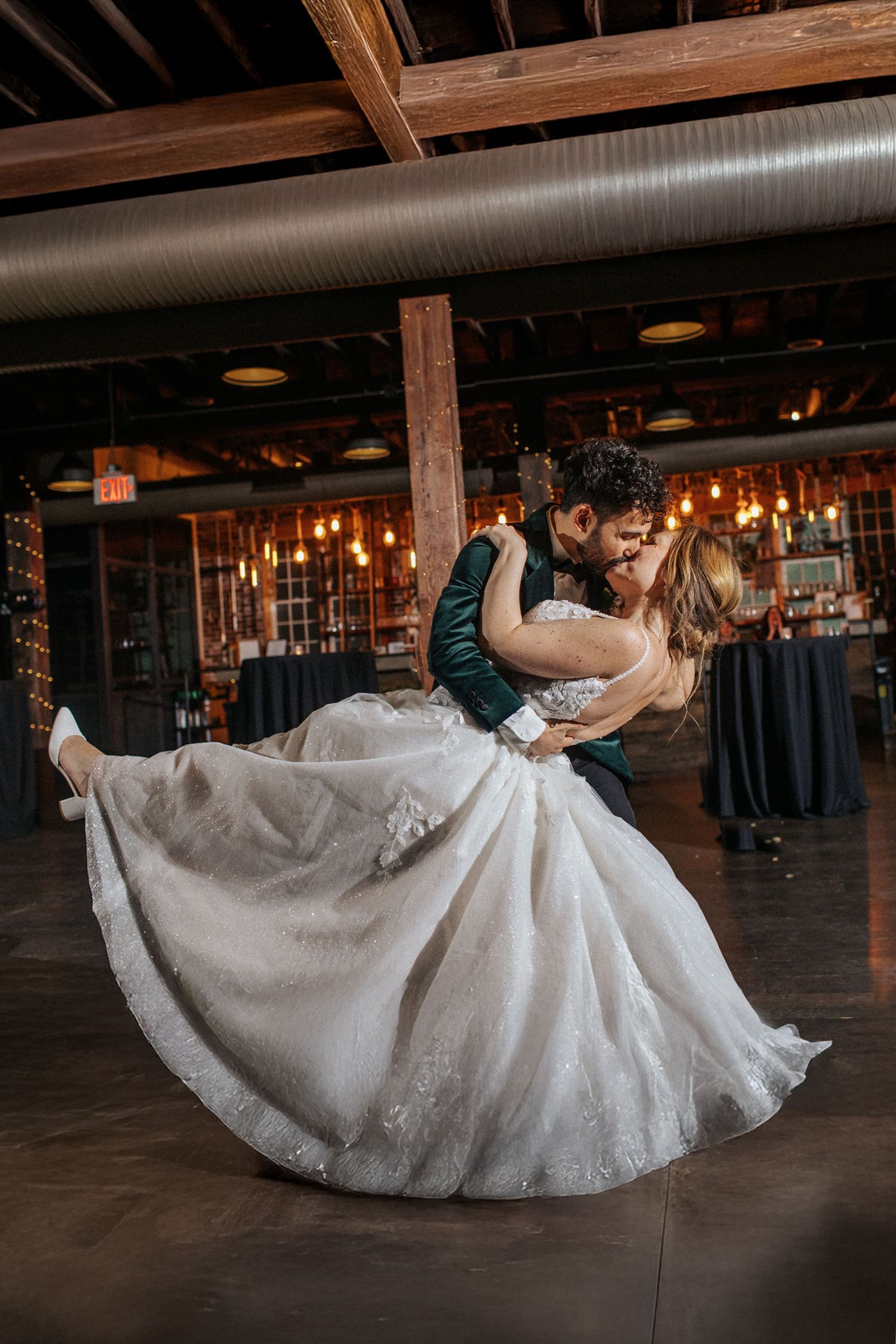 A dramatic dance dip shows a bride's white dress fanning out across a wooden floor during a wedding reception.