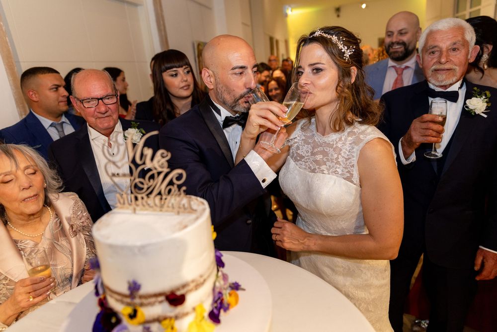 Bride and groom sipping champagne together at wedding reception with white cake and guests in background.