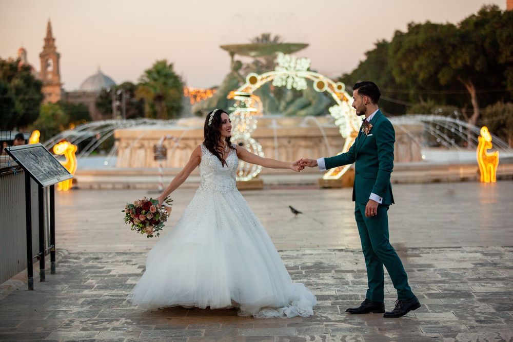 Bride and groom hold hands near an illuminated fountain at dusk in a scenic plaza during their wedding celebration.