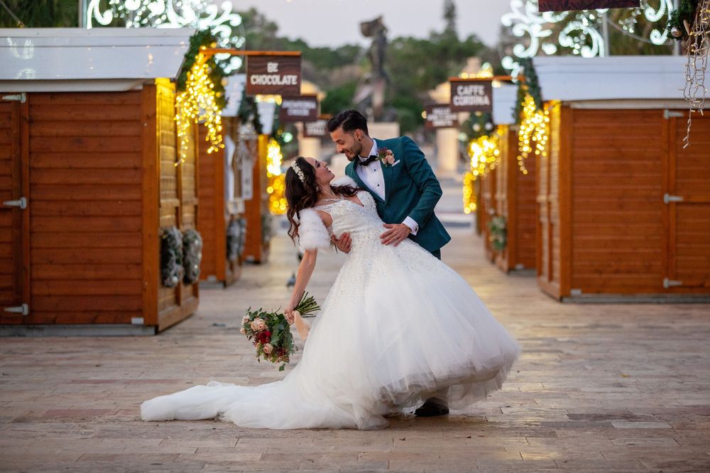 Bride and groom share a romantic dip kiss on a festive outdoor pathway lined with wooden booths and warm string lights.