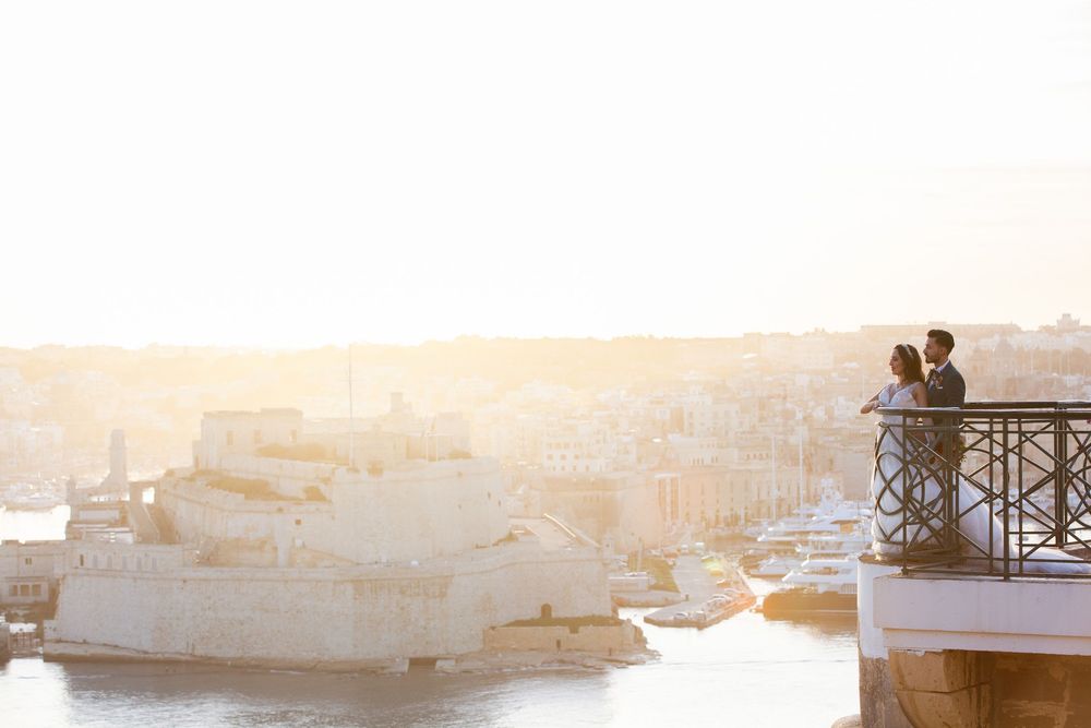 Couple sharing a romantic moment on a sunlit balcony overlooking Malta's Grand Harbour at golden hour.