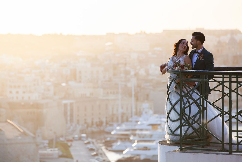 Newlyweds embracing on a balcony overlooking a sunlit Mediterranean cityscape at sunset.