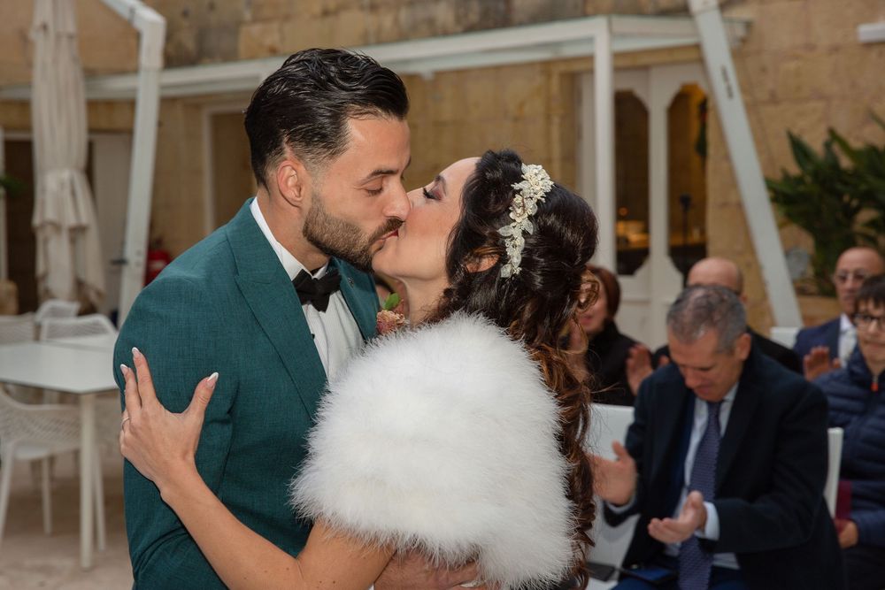 Newlywed couple sharing a romantic first kiss, groom in green tuxedo, bride in white fur stole at wedding ceremony.