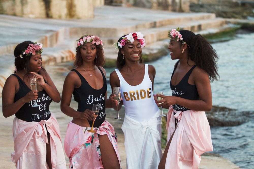 Four women in bridal party swimsuits and floral crowns pose by the water during a bachelorette celebration.