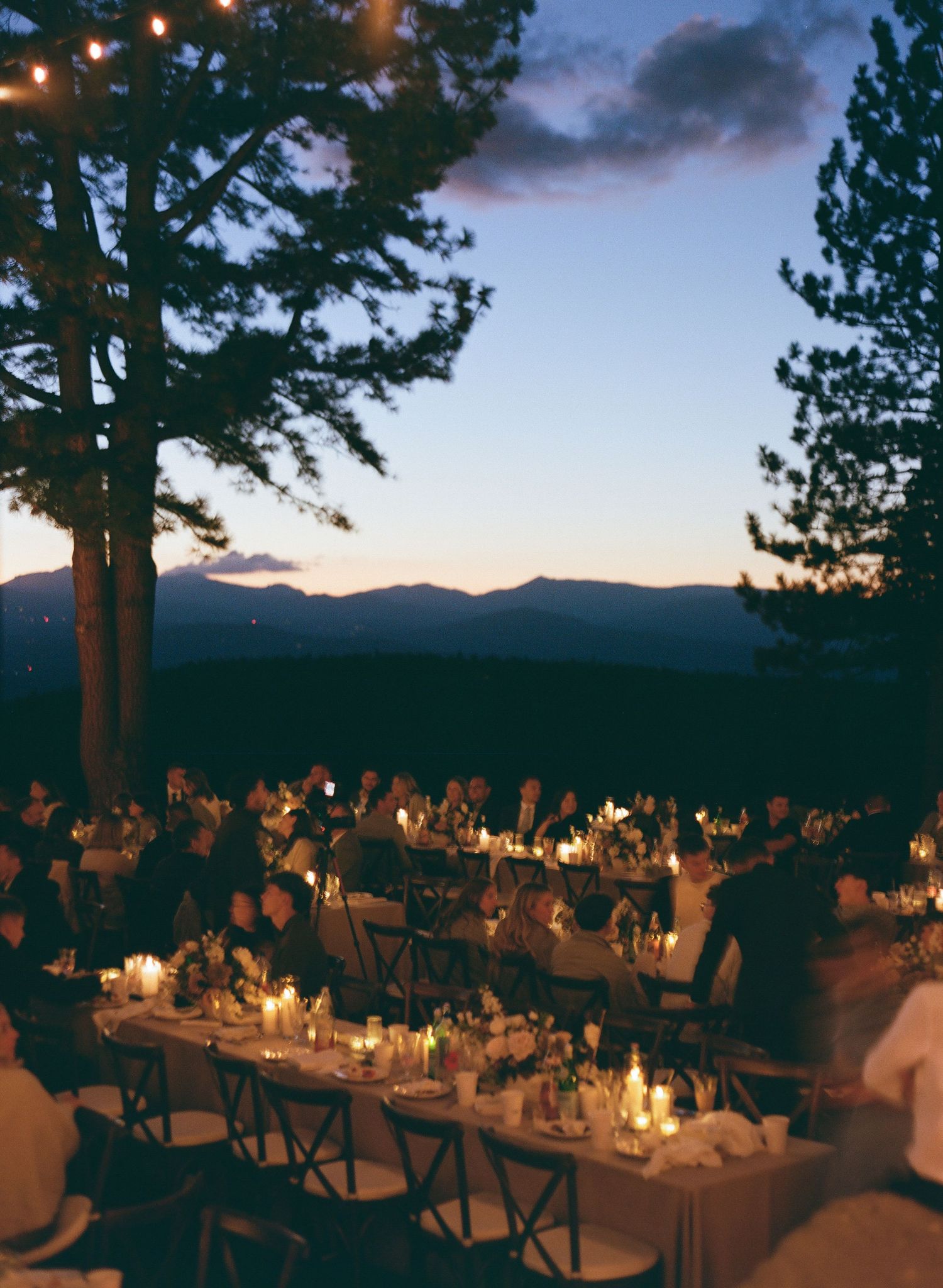 An outdoor wedding reception at dusk with candlelit tables and pine trees silhouetted against a dramatic mountain sunset.