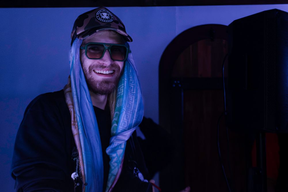 DJ wearing sunglasses and headphones performs at a dimly lit venue with blue atmospheric lighting.