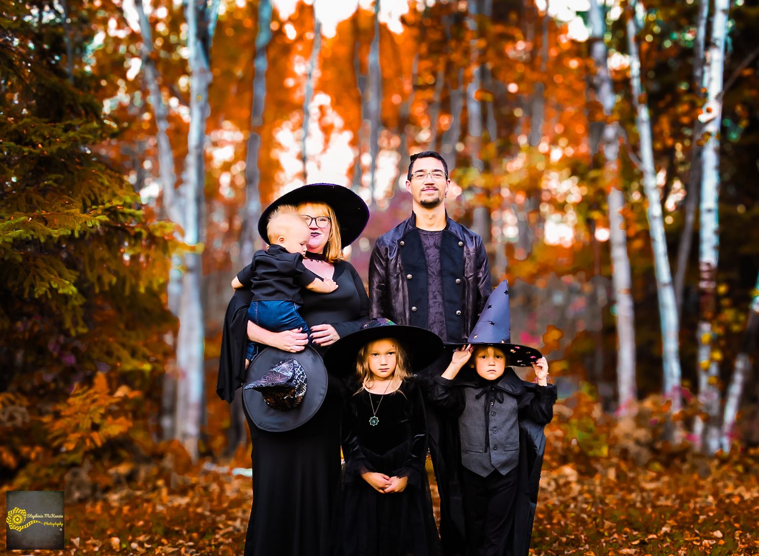 A family wearing dark clothing poses for autumn portraits in a forest with vibrant orange fall foliage.