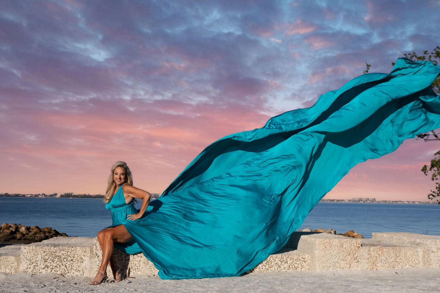 A flowing turquoise fabric billows dramatically against a vibrant sunset sky on a beach.