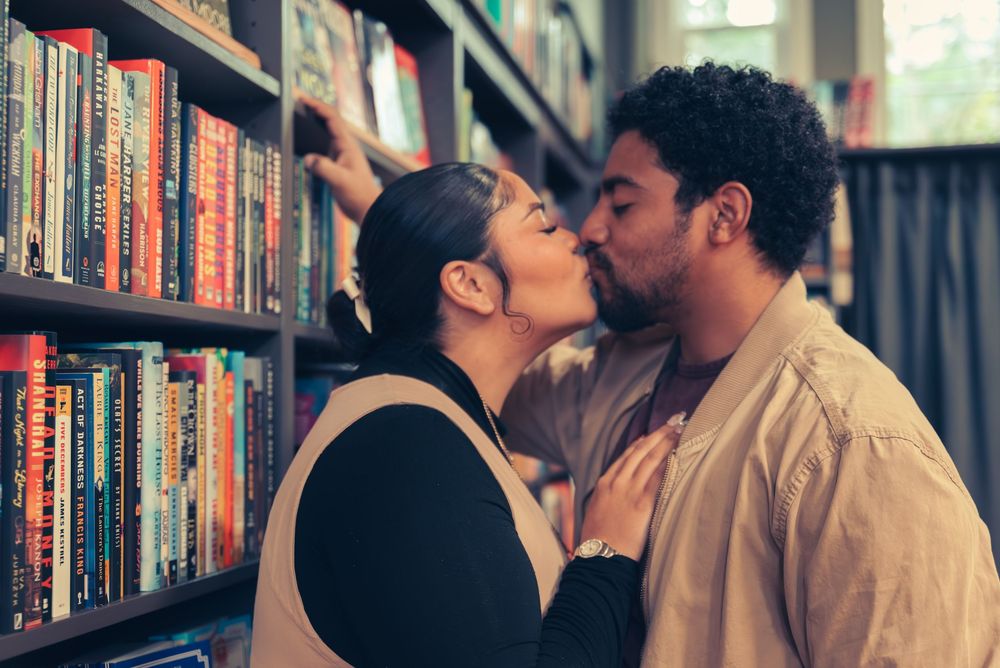 A romantic couple shares an intimate moment in front of a library bookshelf filled with colorful books.